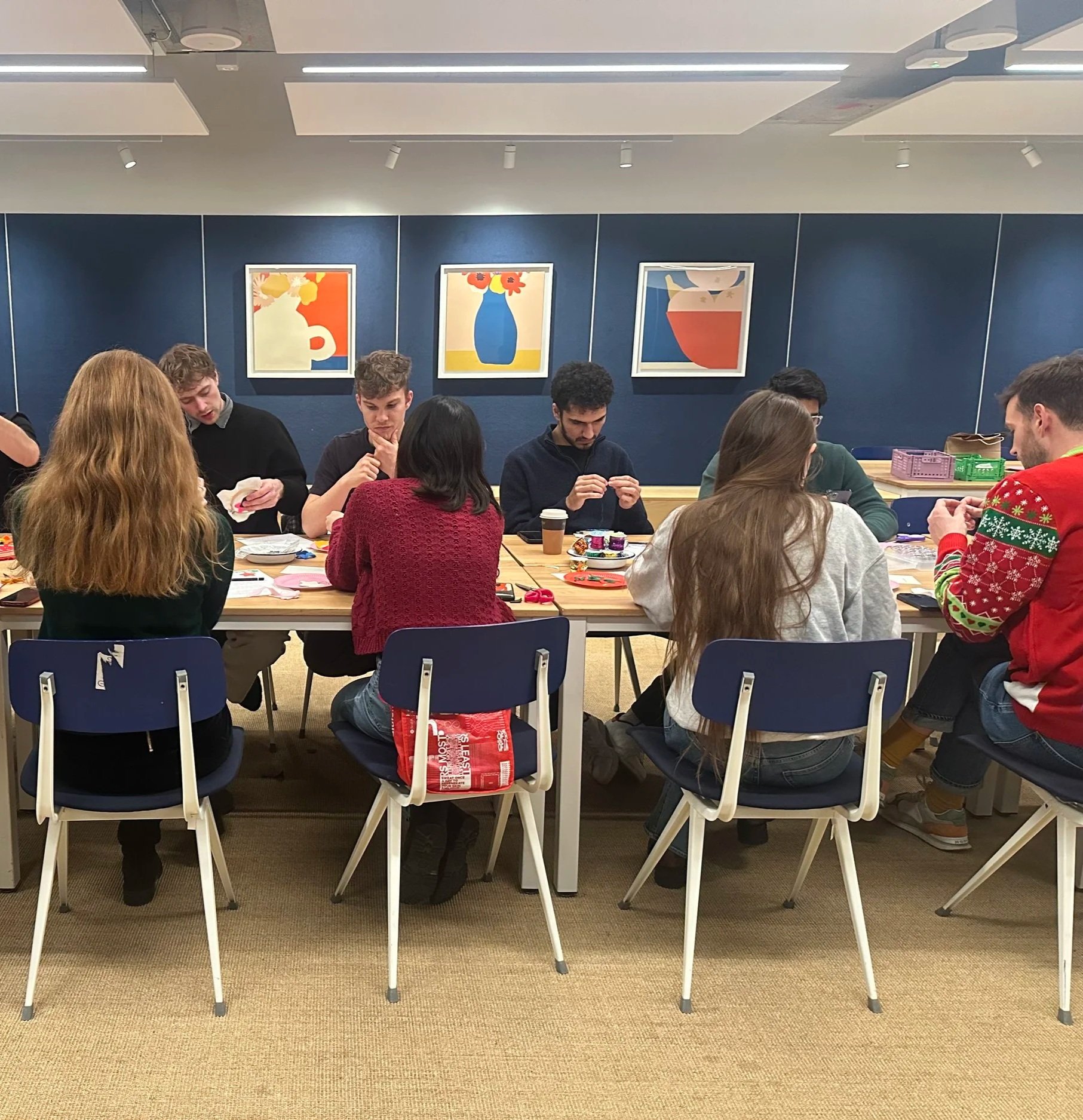 People gathered around a table, engaging in a team building office craft activity, with artwork of colorful vases and flowers on the dark blue wall behind them.