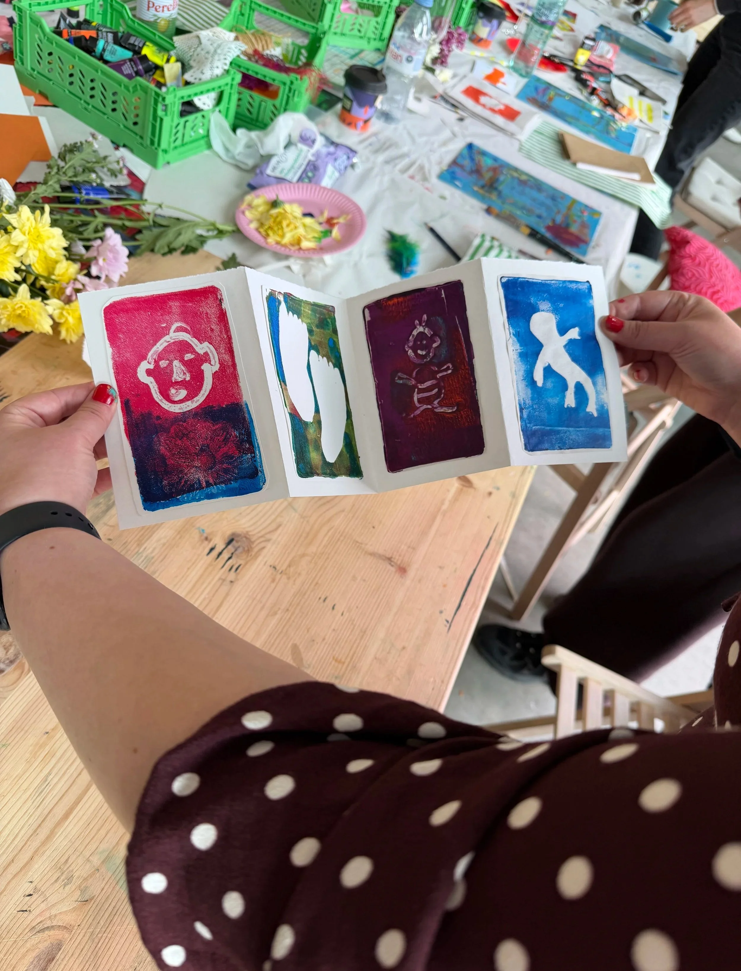 A person holding colorful prints or cards featuring various abstract and expressive designs, with a cluttered table in the background containing art supplies, cups, and paper.