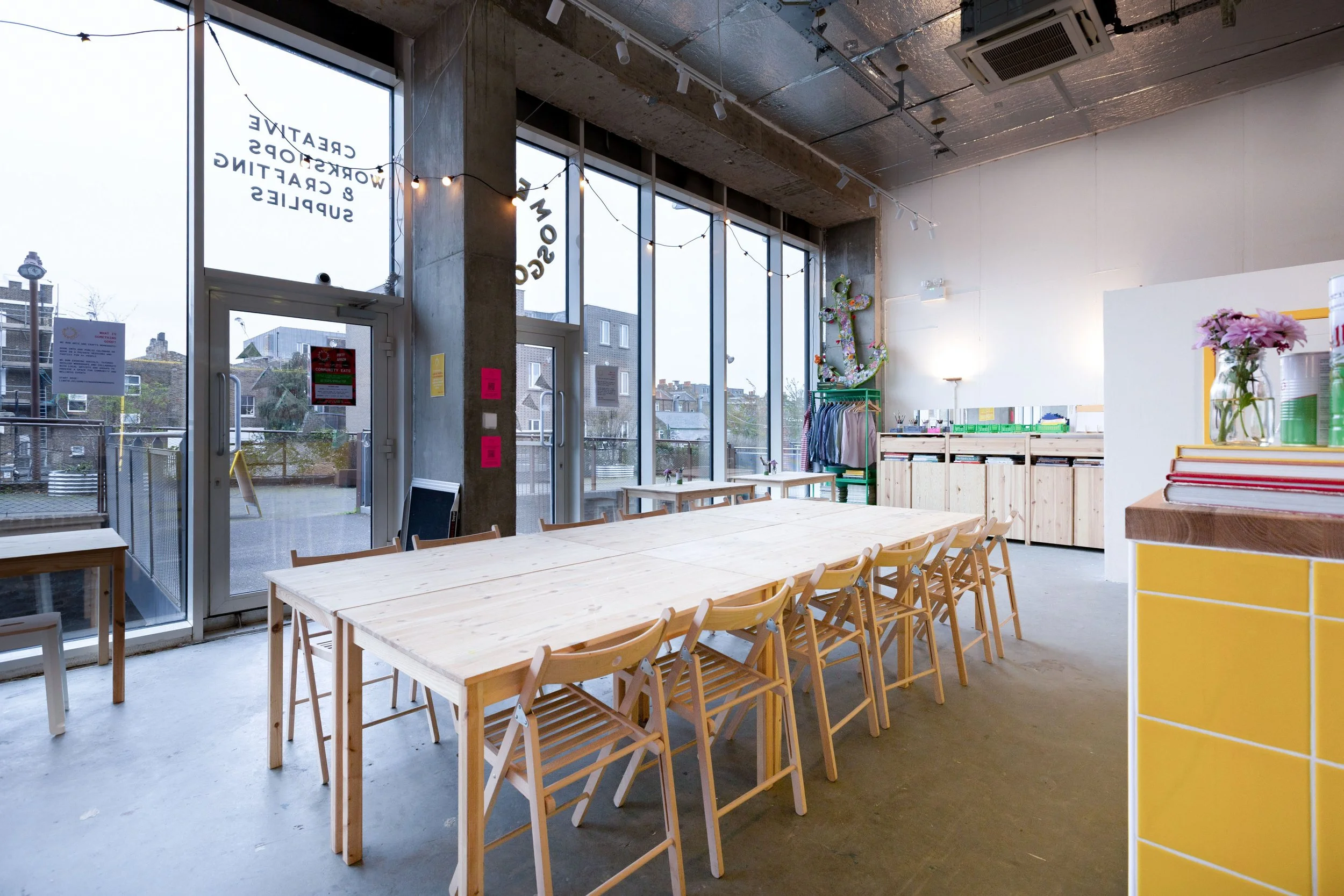 Interior of Something Good Workshops, a modern, minimalistic craft workshop space with large windows, wooden tables and chairs, hanging string lights, and floral decorations.