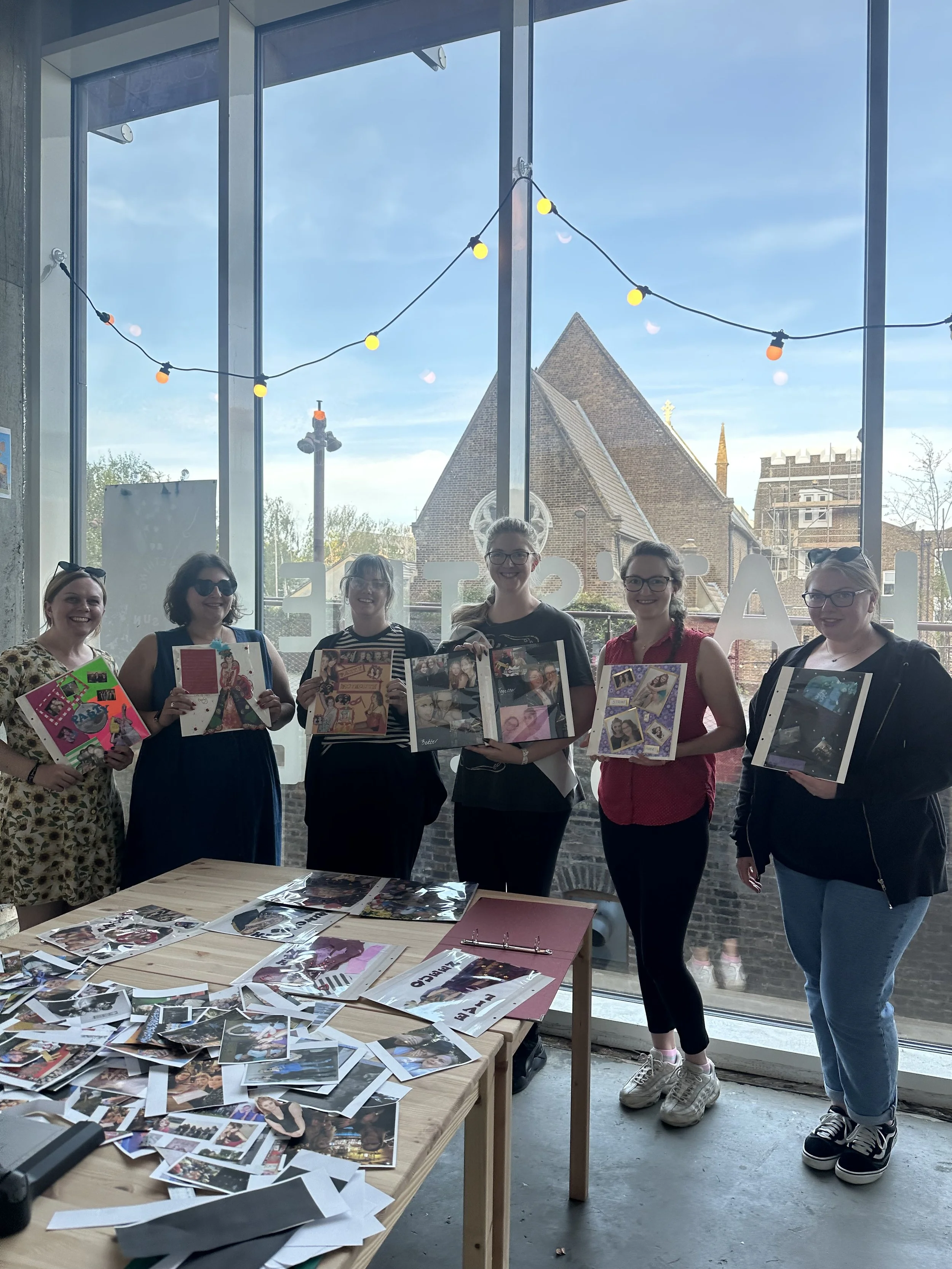 Six women standing inside a room holding collaged pages of photo albums made as part of a hen do party workshop in London. The table in the foreground is covered with loose photographs and scrapbook materials.