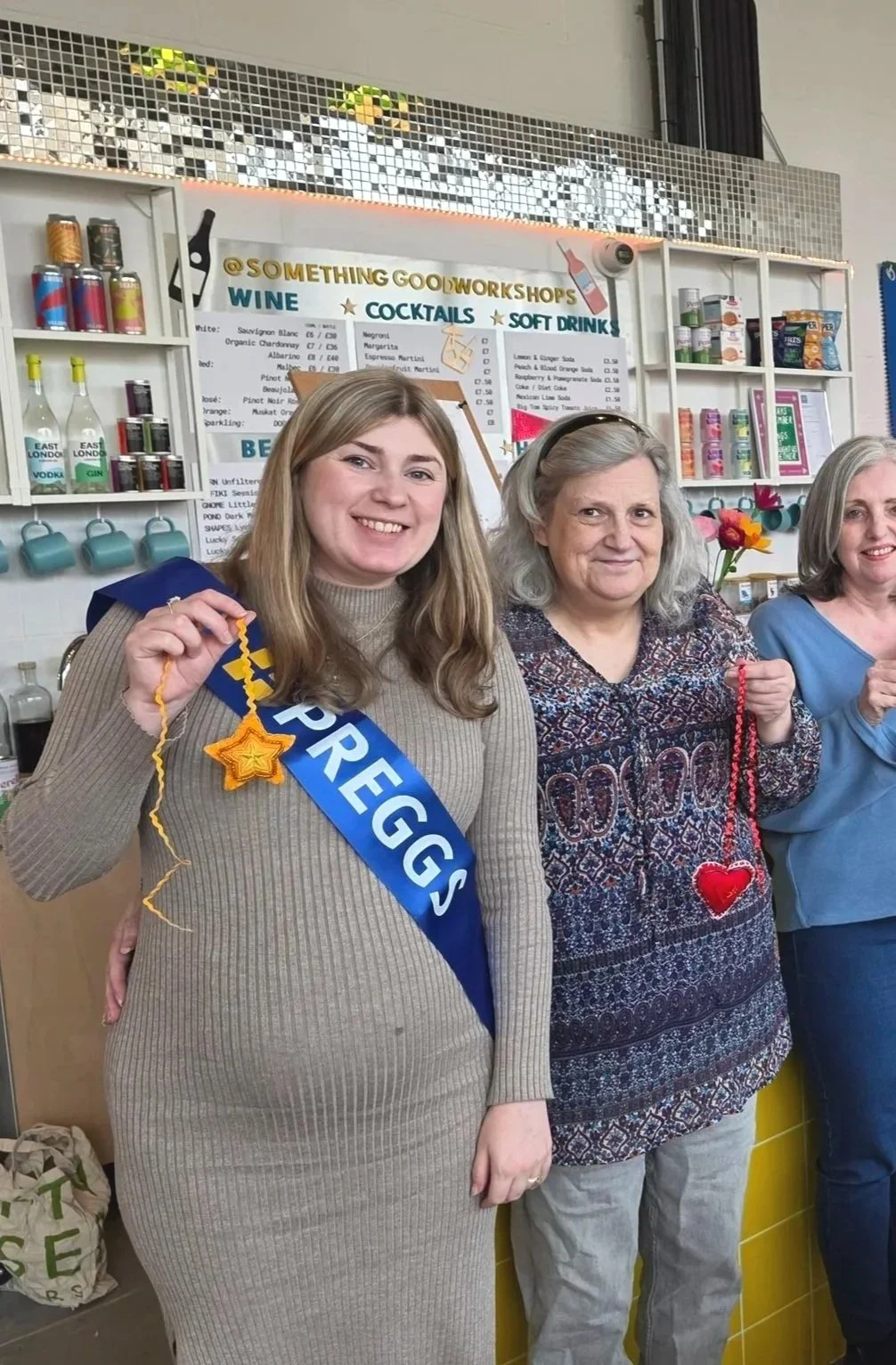 Three women standing in a shop, the woman on the left is smiling and wearing a 'PREGNANT' sash, holding a star-shaped decoration. The middle woman is holding a heart-shaped decoration, and the woman on the right is smiling. The shop behind them has a menu and various canned drinks on display.