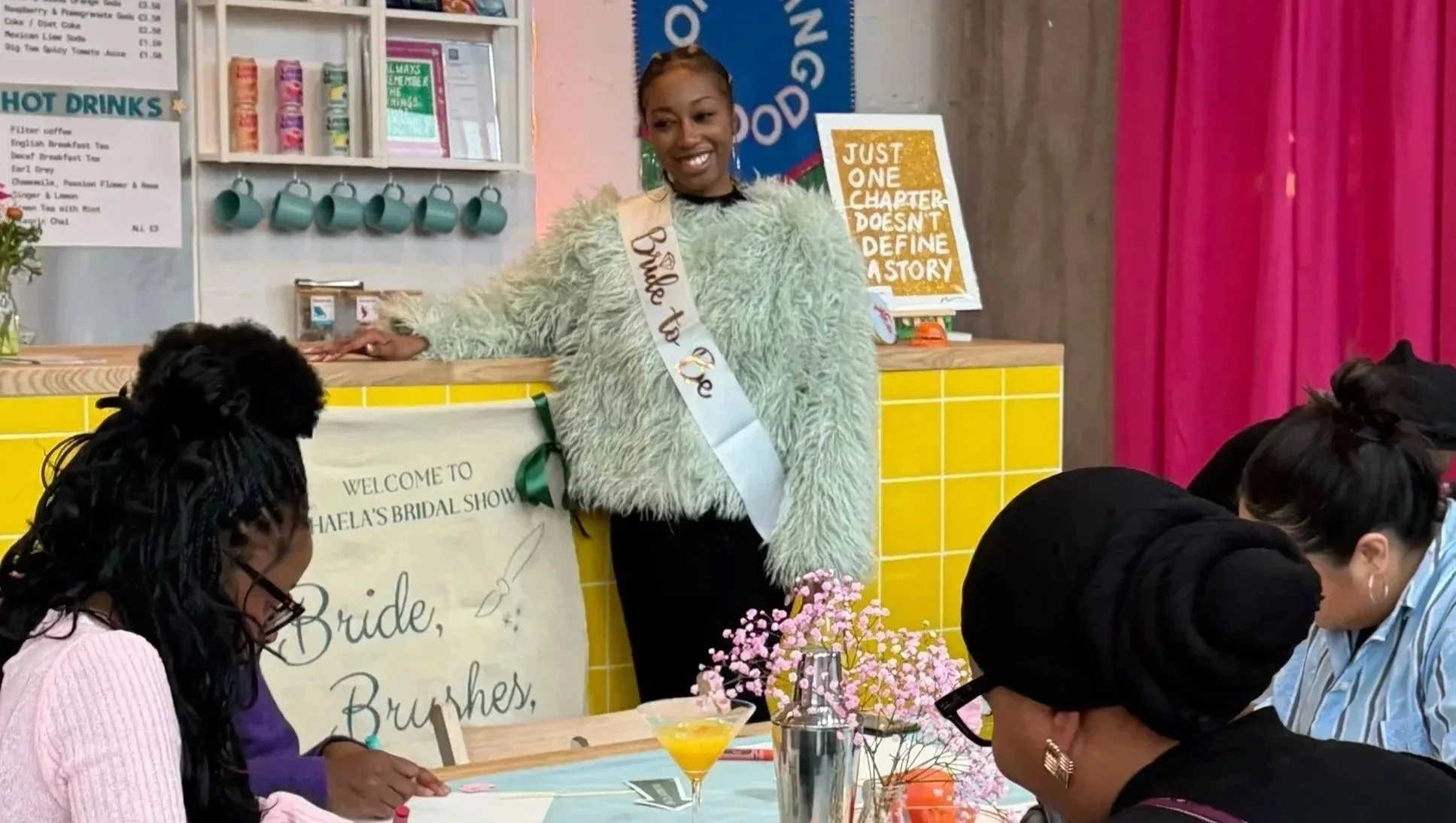 A woman wearing a large green furry jacket and a sash that says 'Bride To Be' is standing behind a table at a bridal event. There are women seated at the table with pink flowers and drinks, and a sign that reads 'Welcome to Mahela's Bridal Show.' Behind her, there is a yellow tiled counter, a poster with a quote about chapters and stories, and a pink curtain.