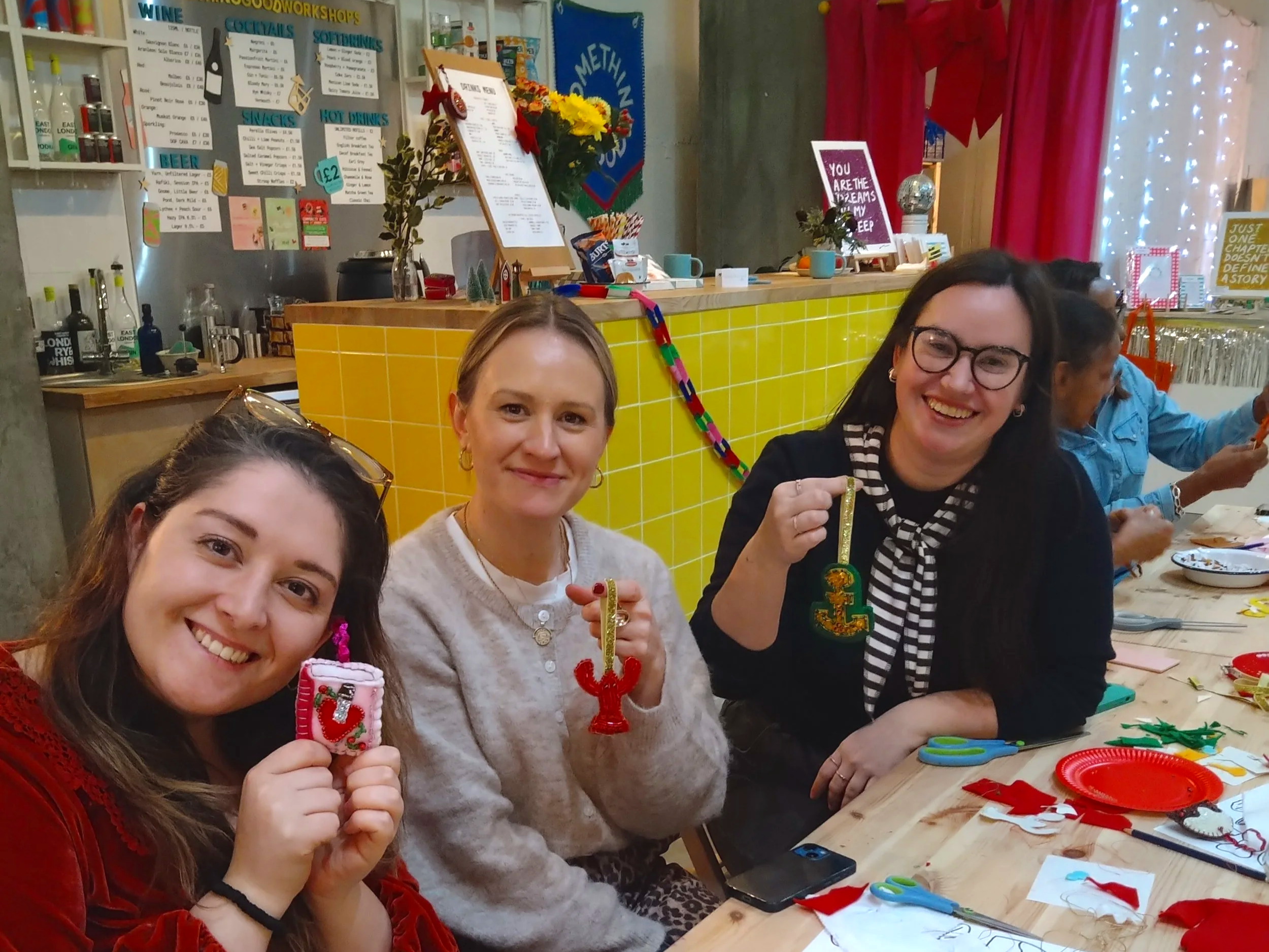 Three women smiling and holding Christmas-themed ornaments at a craft table at Christmas party workshops in London. The background has a menu, colorful decorations, and a yellow tiled counter.