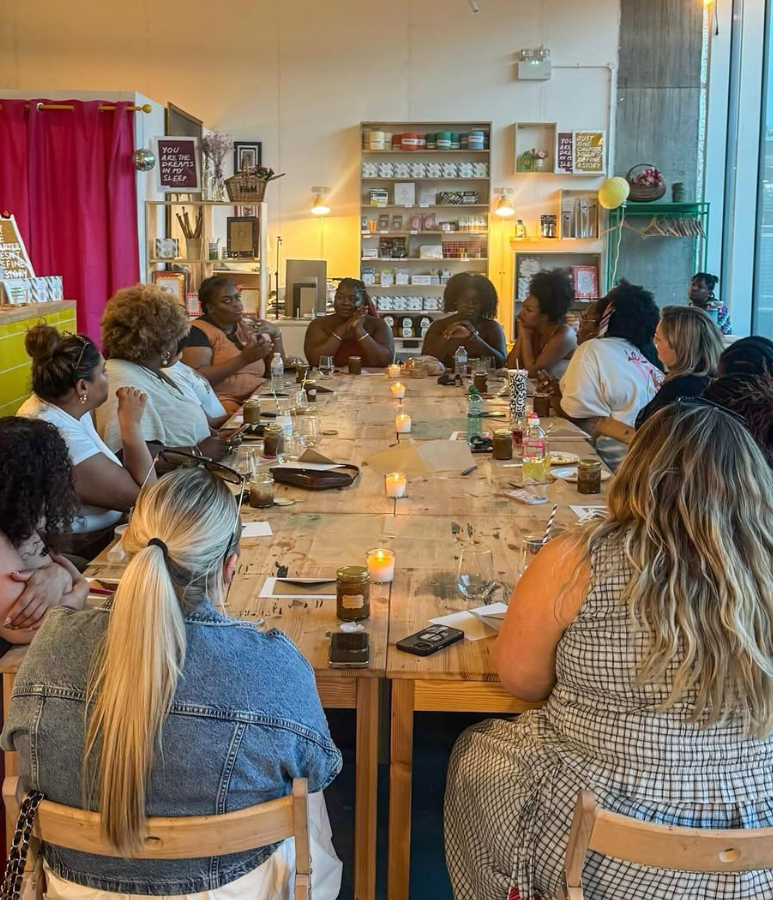 A group of women sitting around a large wooden table in a cozy cafe, engaged in conversation. The cafe has warm lighting, shelves with decorative items, and a window with sunlight streaming in.