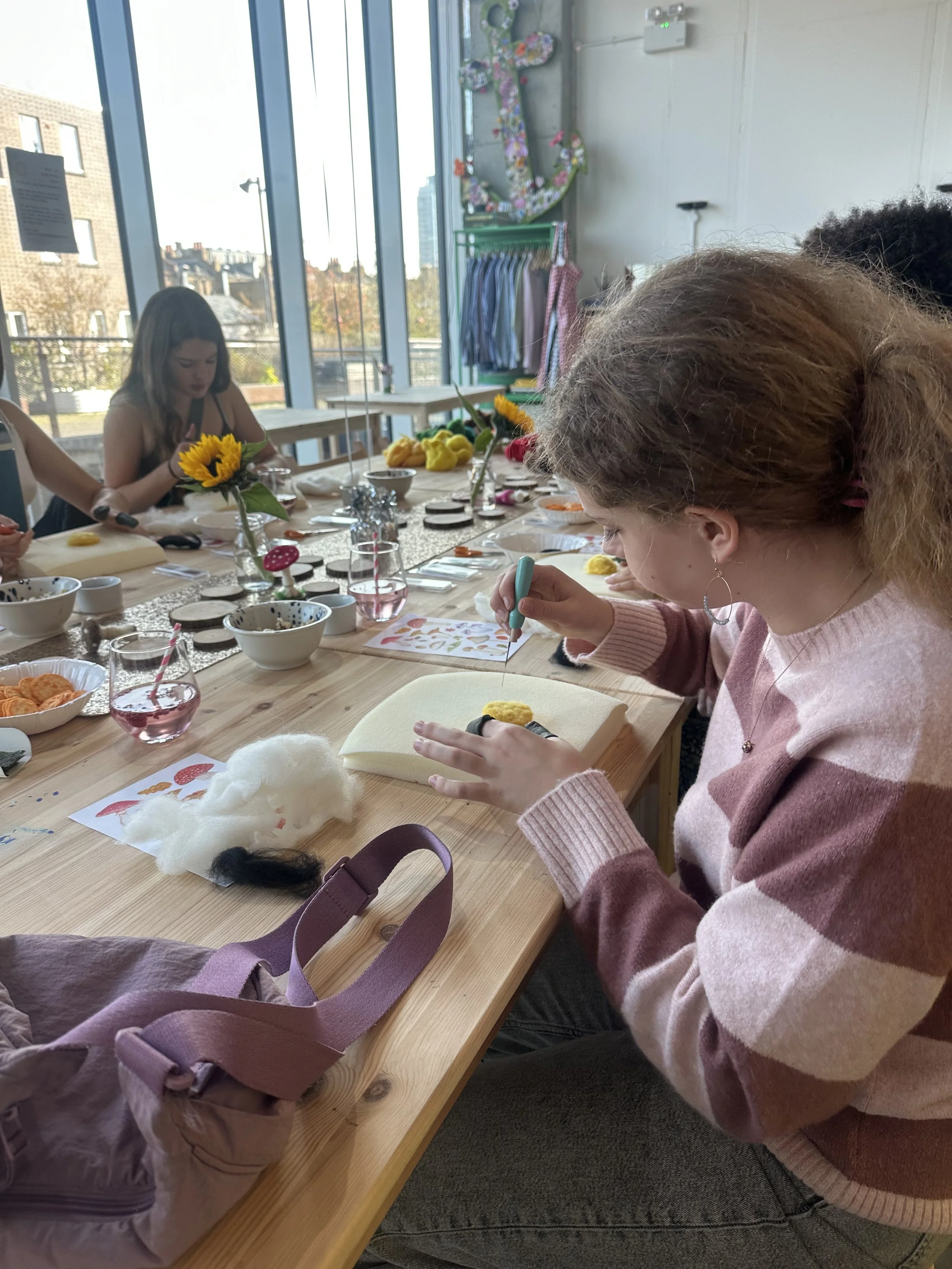 People sitting at a table engaged in arts and crafts activities, with supplies like fabric, paper, and tools, in a bright room with large windows and decorations on the walls at Something Good Workshops in Deptford, London. 