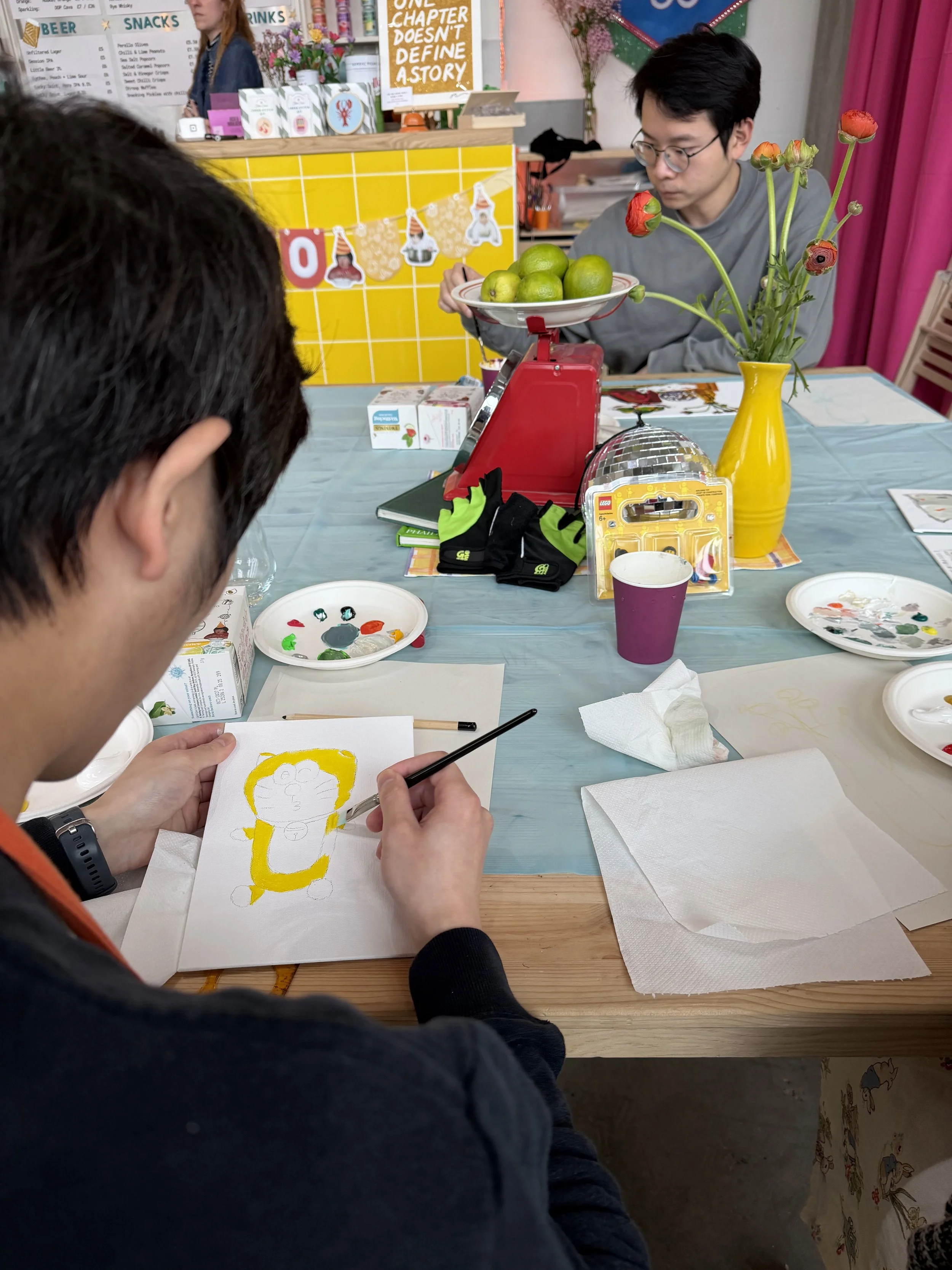 A person is painting a cartoon lion on paper with yellow paint, sitting at a table with art supplies, tissues, and a paper plate.