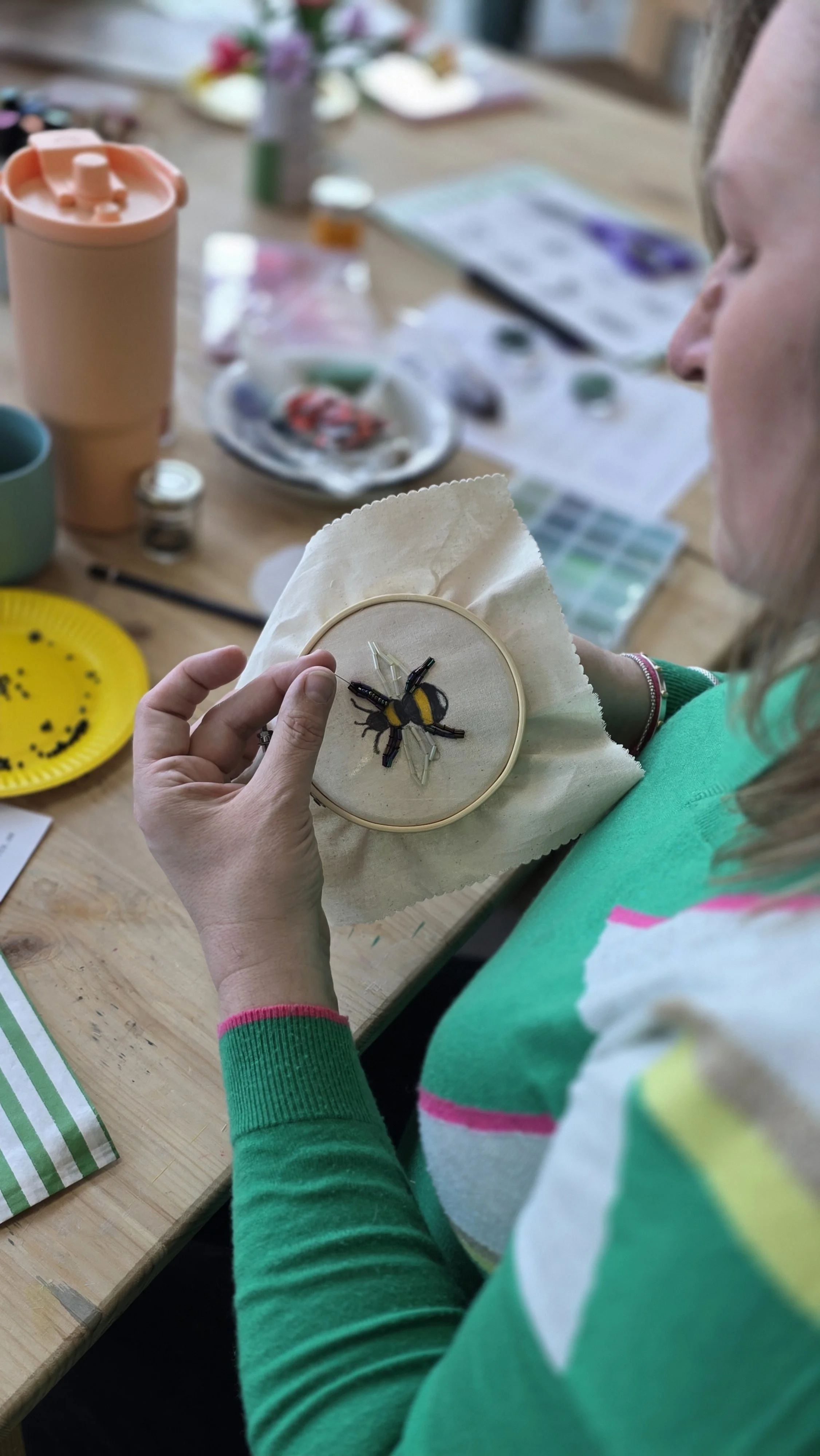 Person holding a bee craft in an embroidery hoop, surrounded by art supplies on a wooden table and a beading and embroidery workshop at Something Good Workshops in Deptford. 
