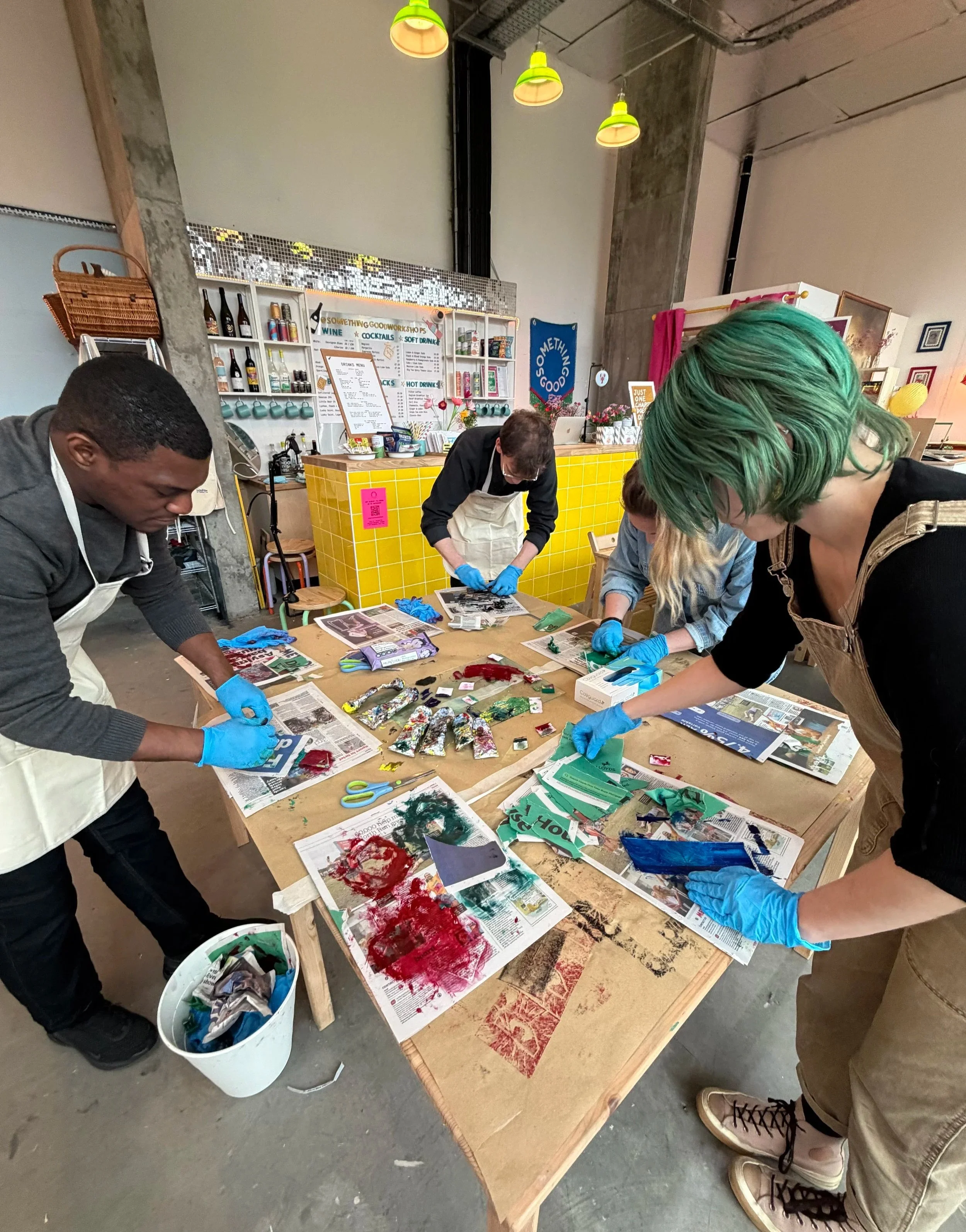 People working on art projects at a table covered with newspapers and various art supplies in a creative studio or workshop space.