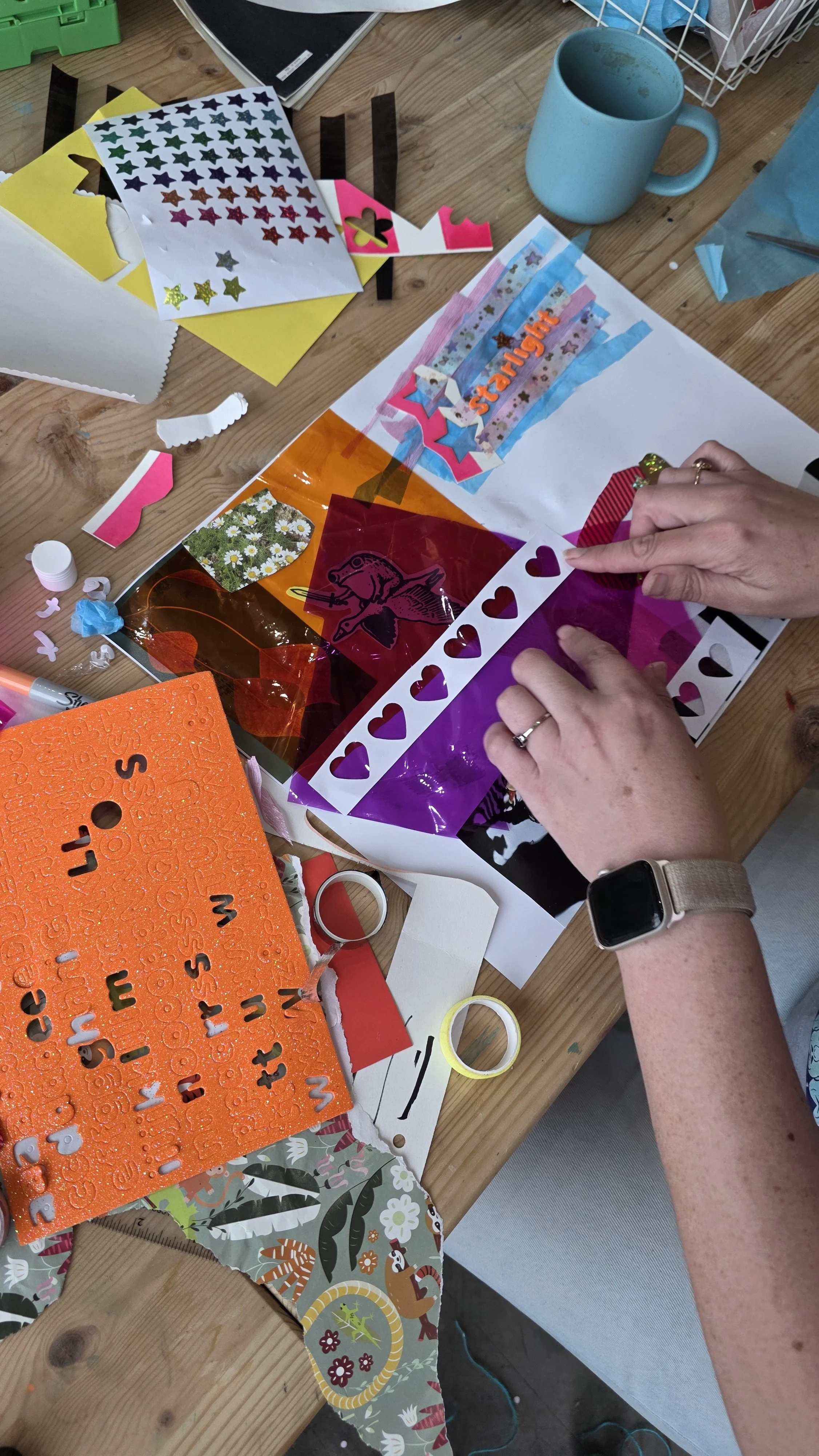 A person's hands working on a colourful collage craft project at a wooden table, surrounded by various craft supplies including stickers, tape, paper, and decorative elements.