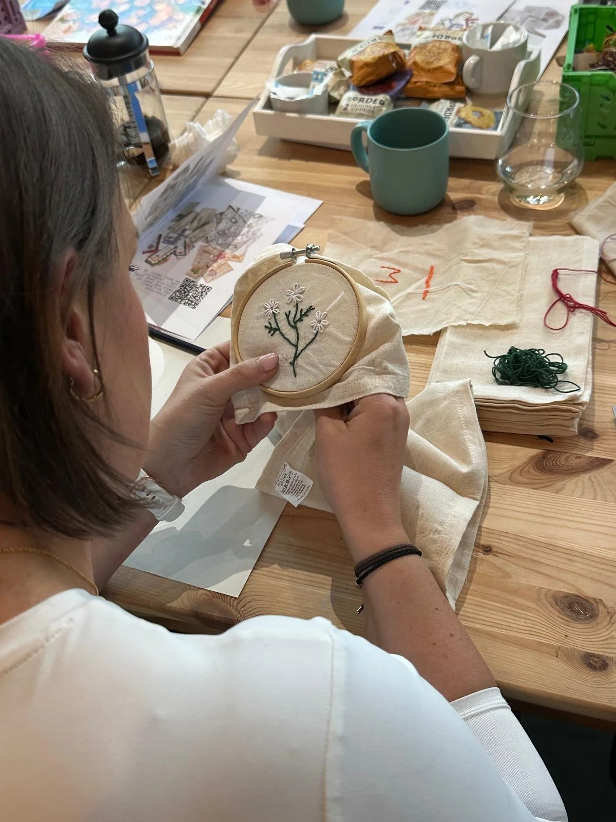 A woman crafts floral embroidery on fabric, sitting at a wooden table with sewing supplies, a cup, and a potted plant.