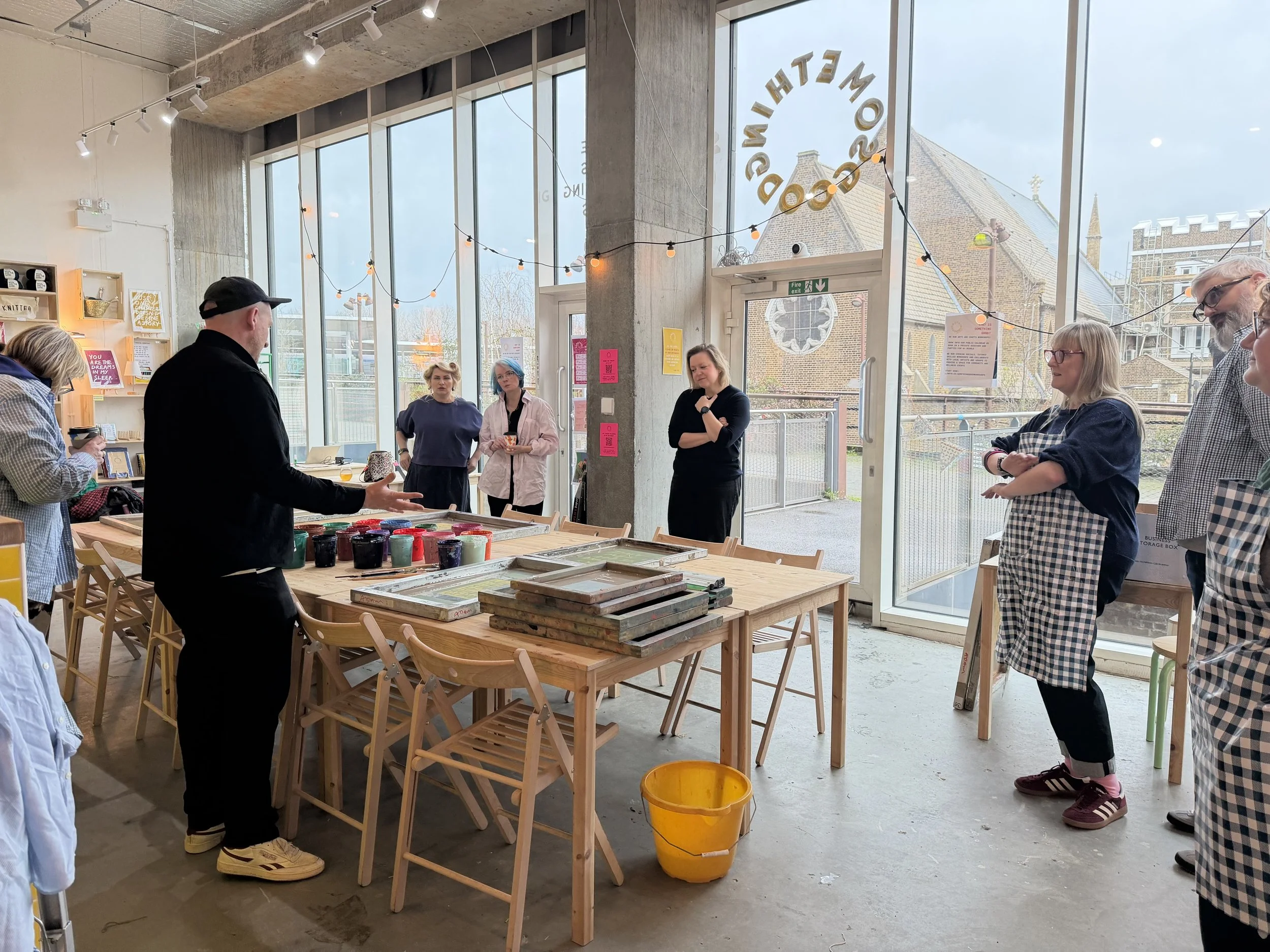 People gathered inside  a modern arts and craft studio, with large windows, string lights, and tables with art supplies. Some individuals are discussing or observing, with a woman in an apron to the right and a man in a black cap in the center.