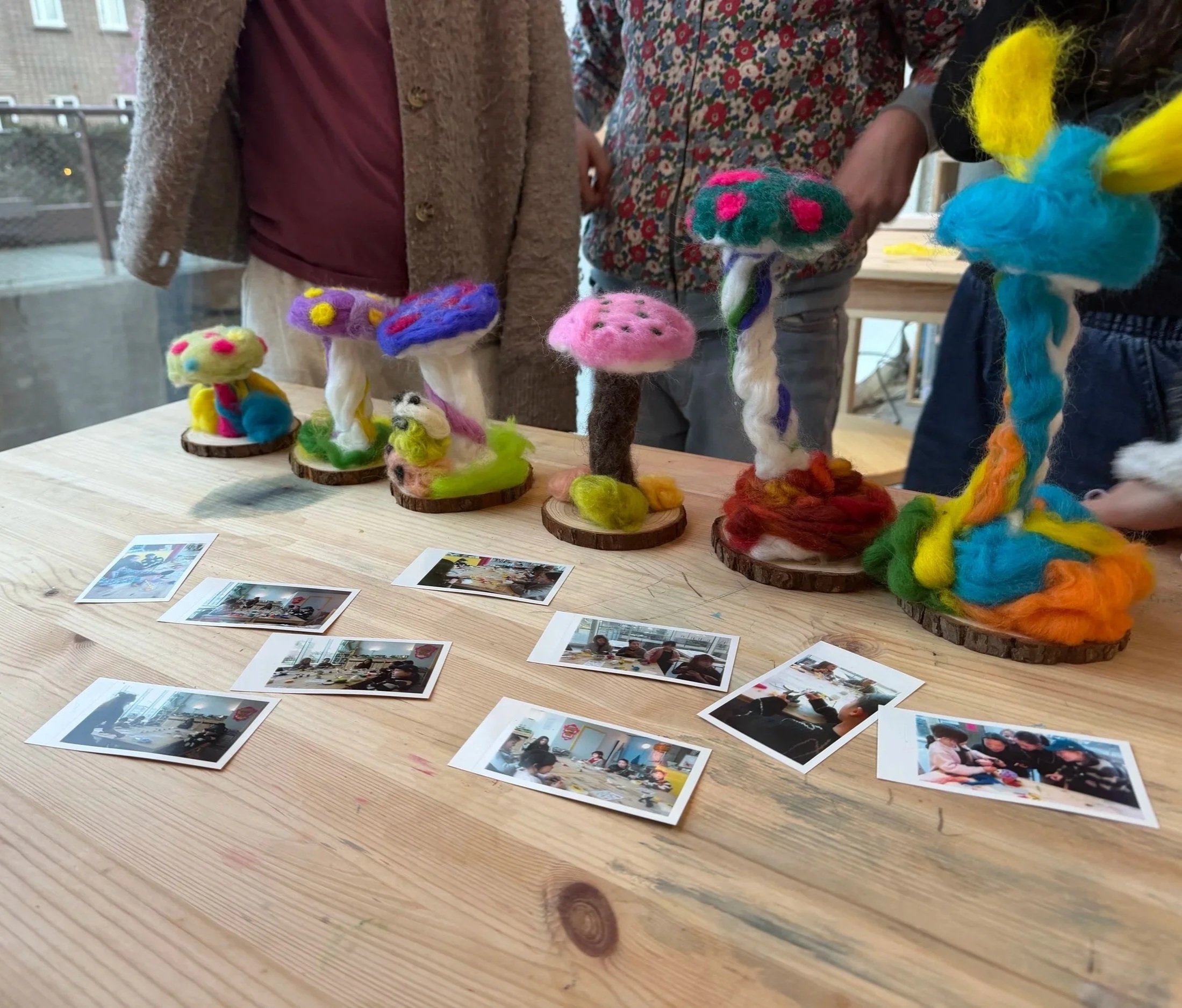 Colorful felted mushroom-like sculptures on wooden bases displayed on a wooden table, with printed photographs spread out nearby, in a room with large windows at a teen birthday party at Something Good Workshops, Deptford, London.