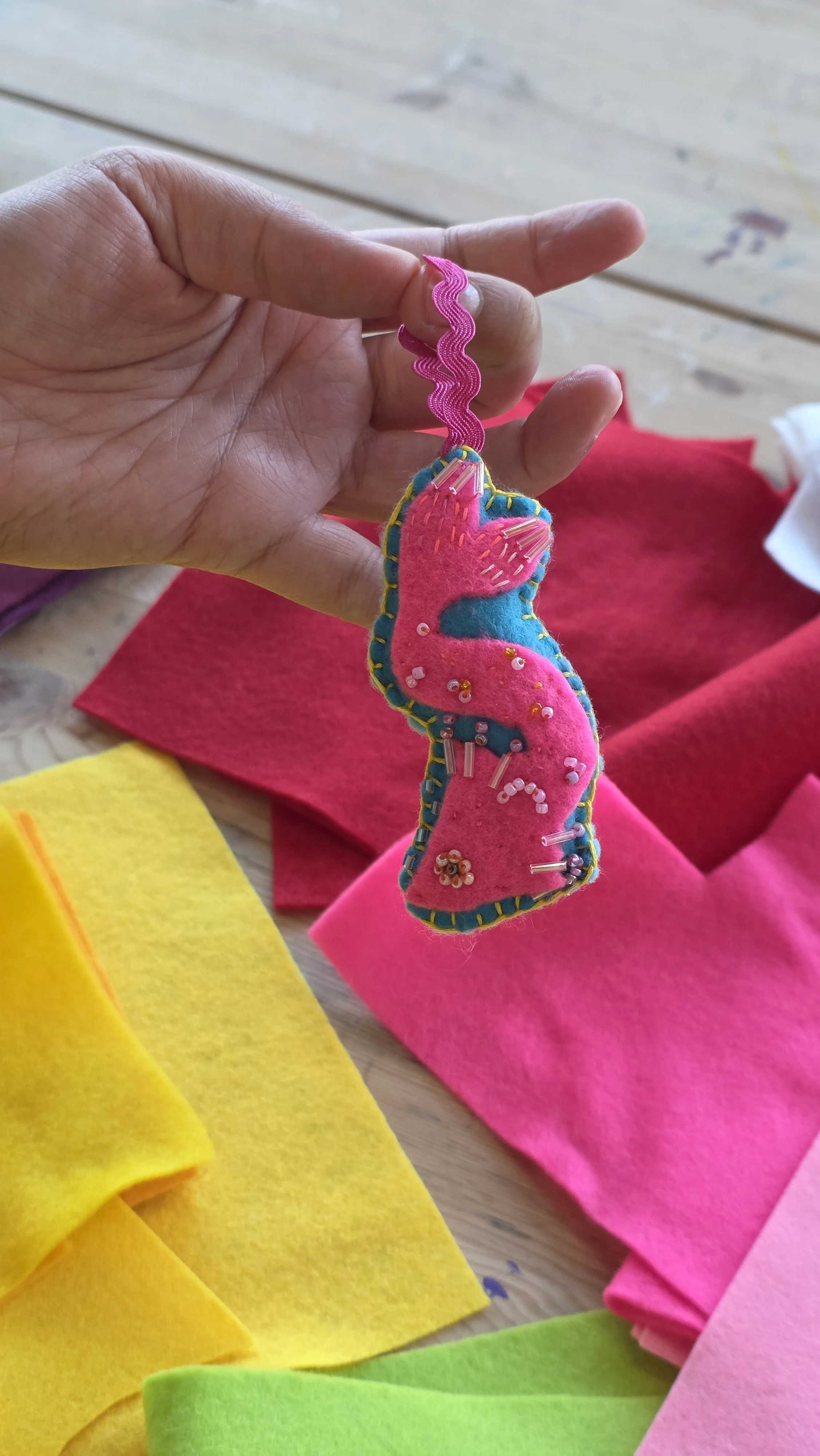 A handmade felt and fabric fish felt charm with pink, blue, and yellow accents, decorated with beads and embroidery, being held by a person's hand.