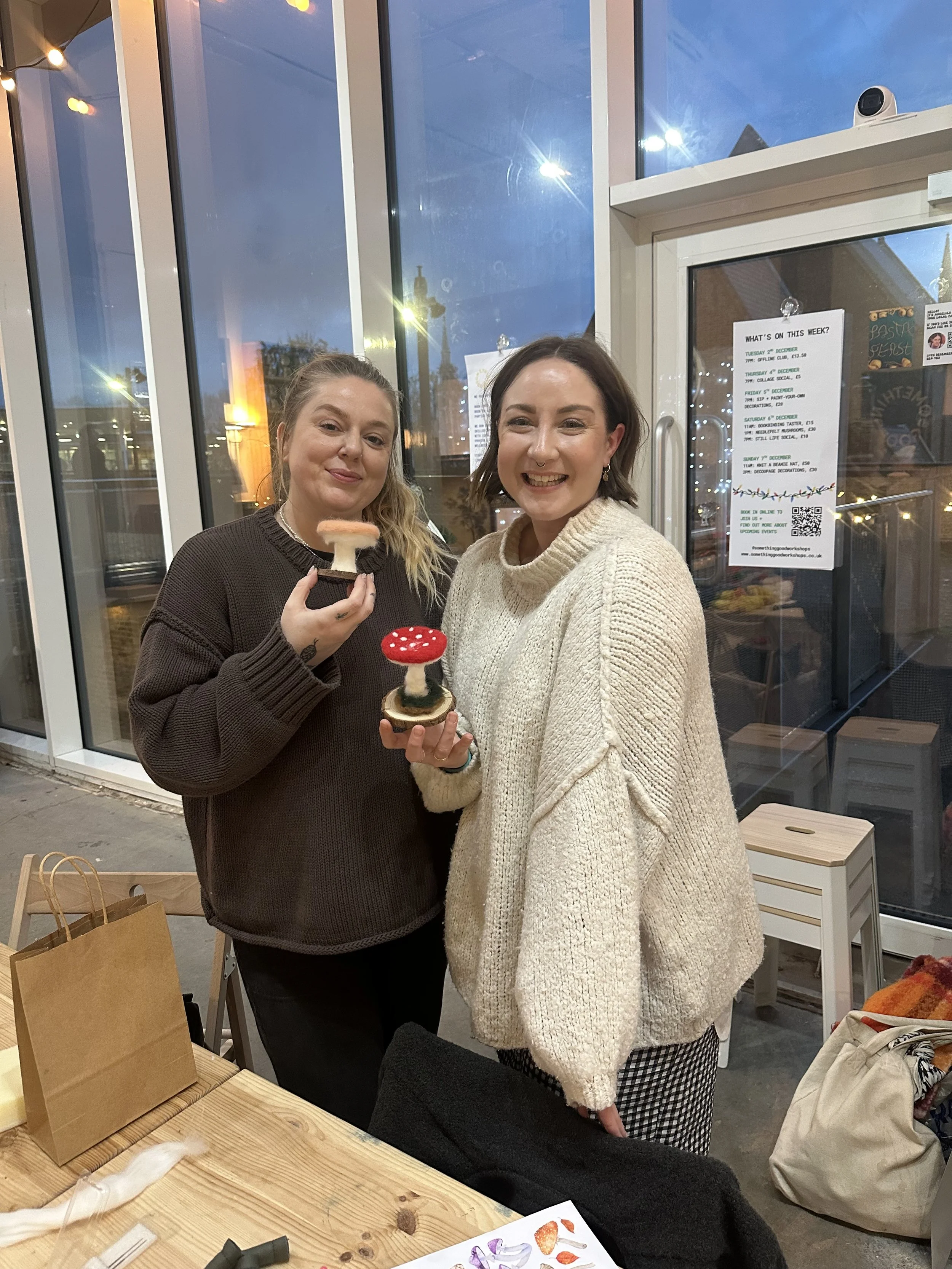 Two women smiling and holding mushroom-shaped sculptures inside a bright, glass-walled room during evening. One woman wears a dark sweater, the other a cream-colored knit sweater. There is a table with a paper bag and some artist supplies in front of them.