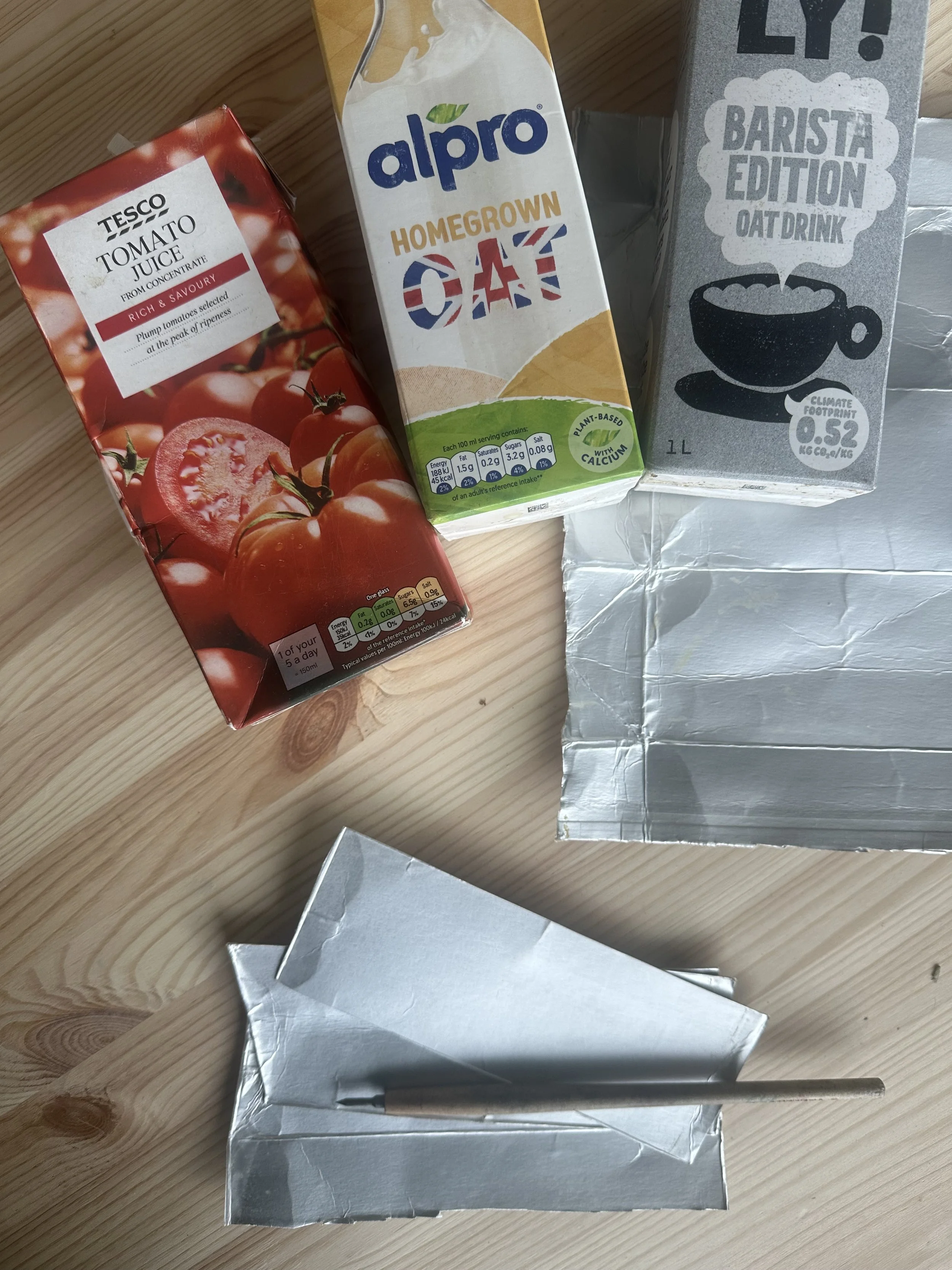 Tea, juice, and oat drink cartons on a wooden surface, with a foil bag and a metal straw in a silver holder.