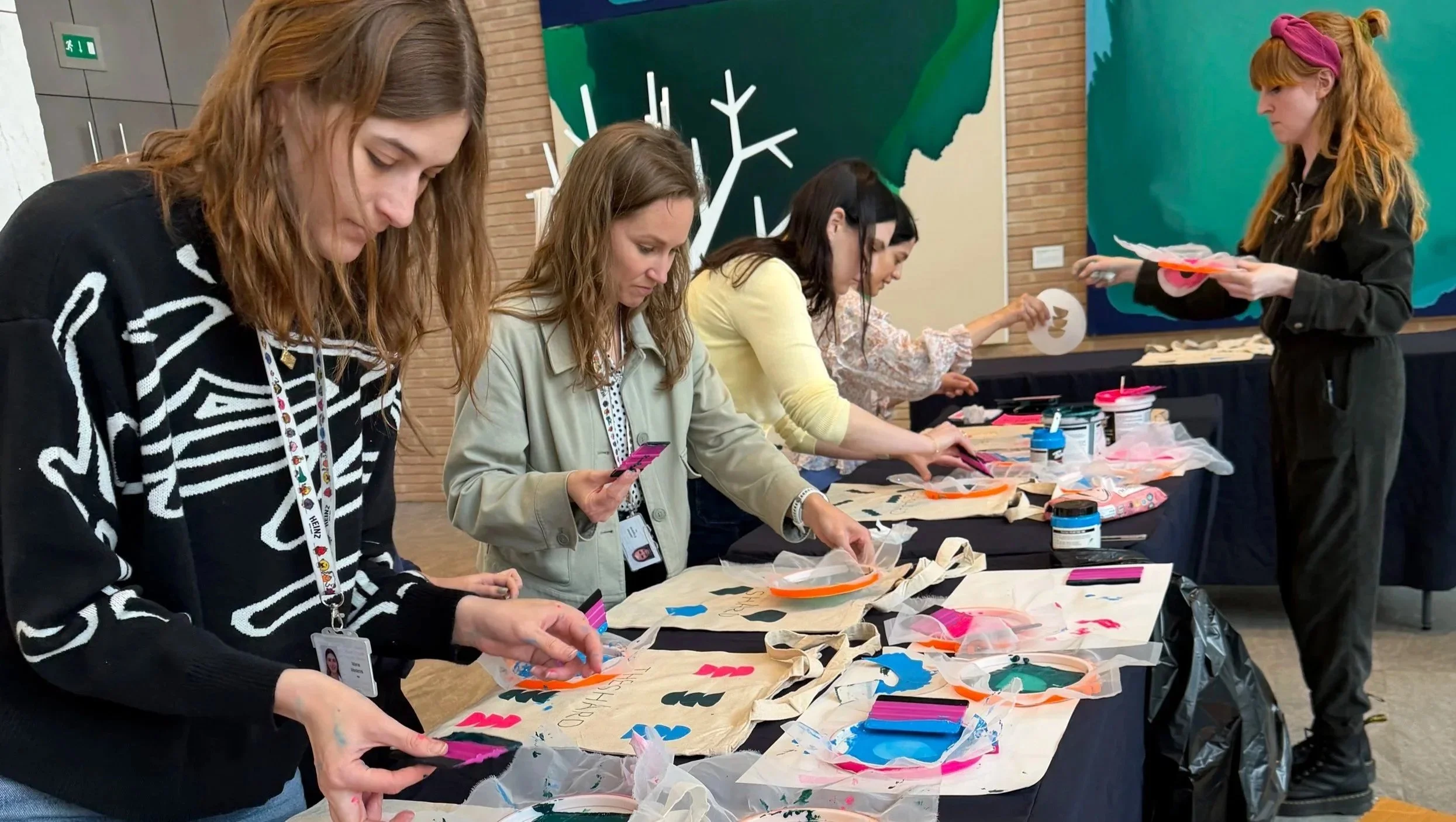 Group of women standing at a table making decorated tote bags with paint and stencils at an indoor event.