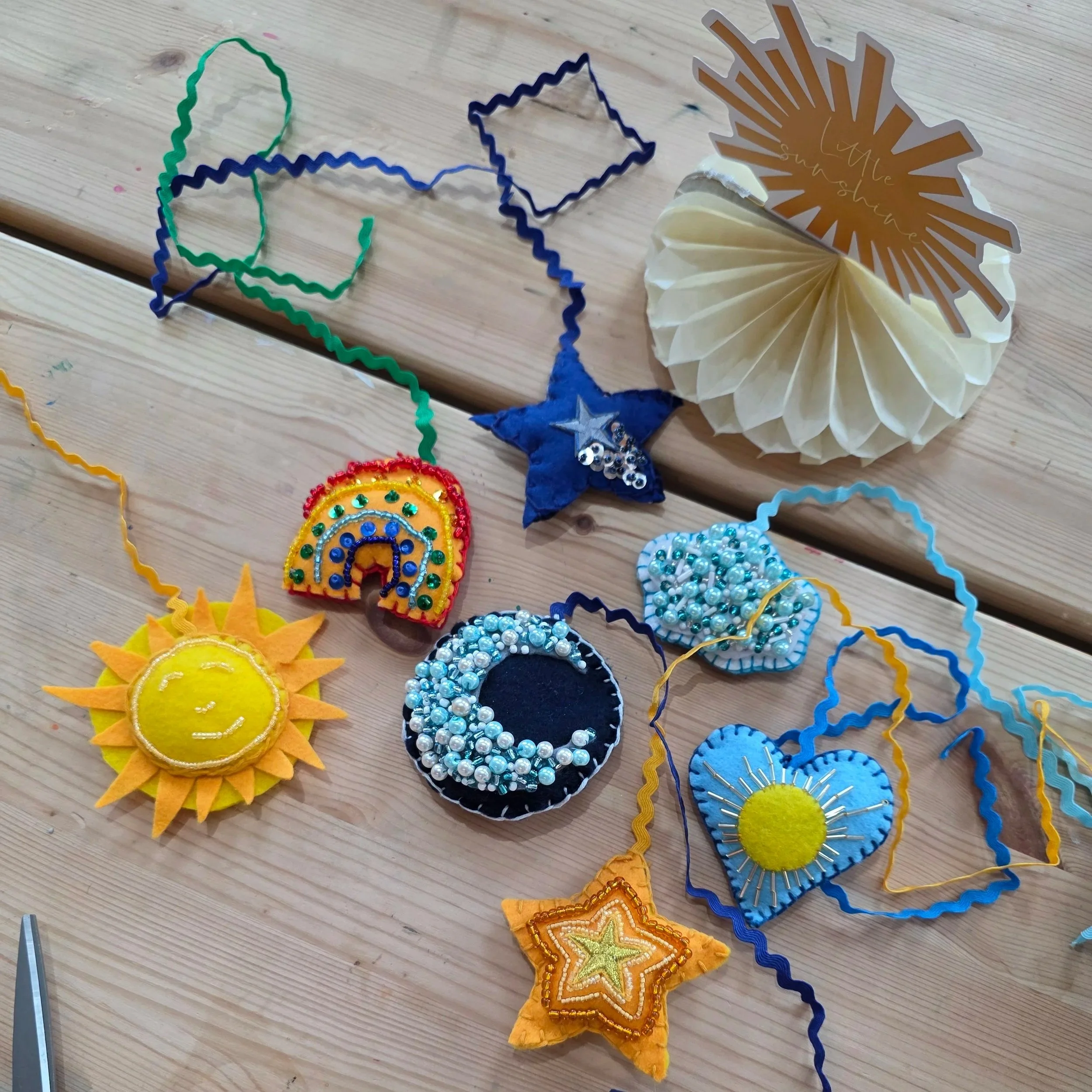 Colorful sun-shaped decorations made of felt with embroidered and beaded details, along with some star-shaped decorations, arranged on a light wooden surface.