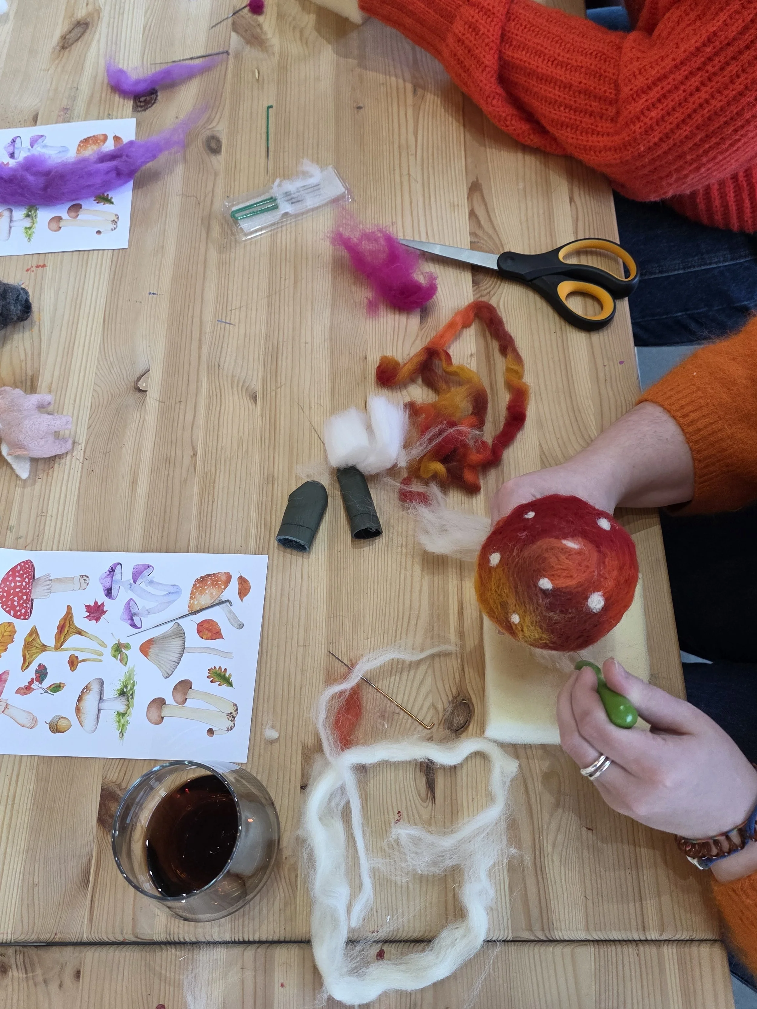 People crafting felt mushrooms and foliage using colored wool, a pair of scissors, and a felting needle on a wooden table. A glass of red wine and an illustrated mushroom chart are also visible.