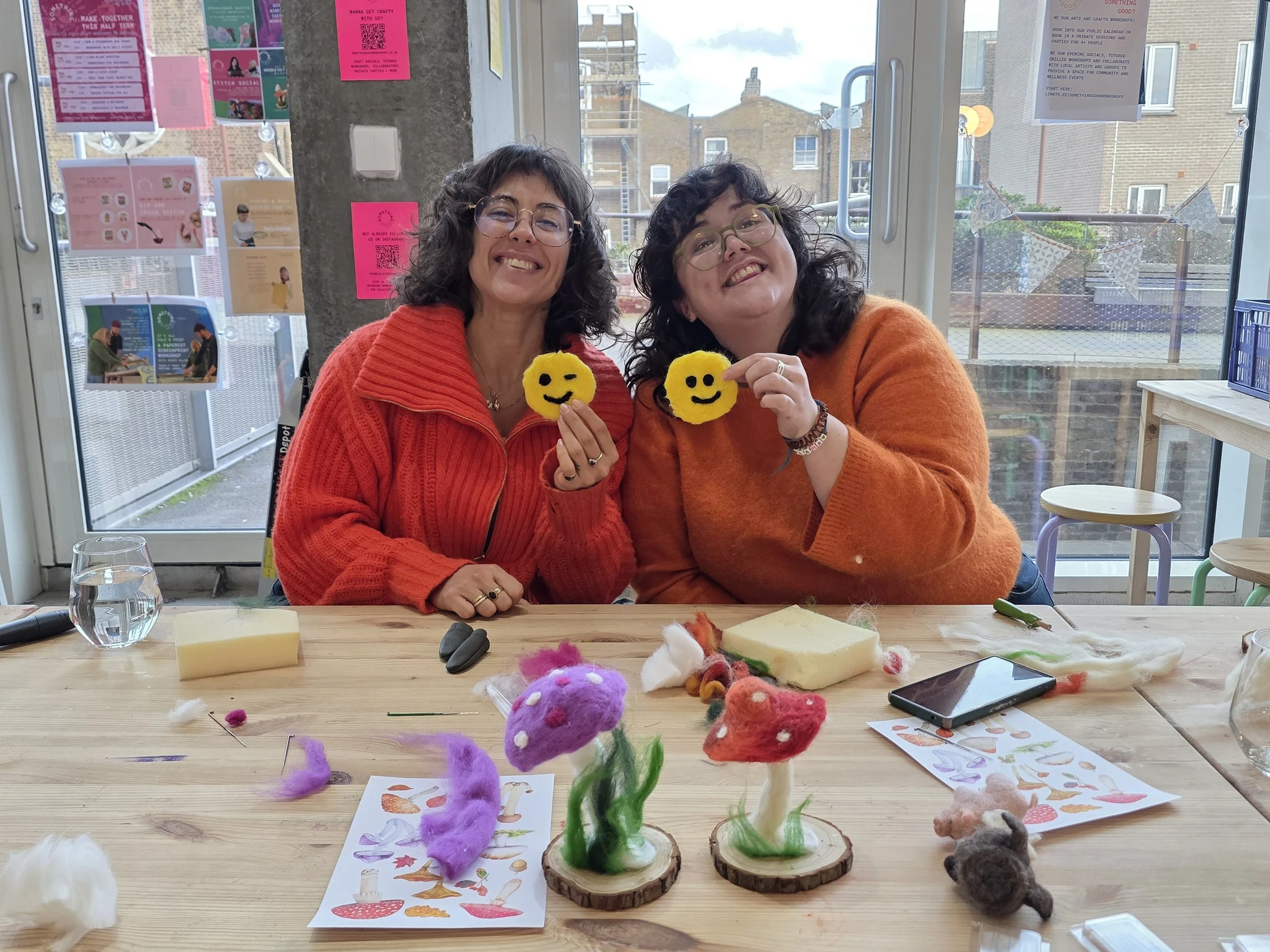 Two women with curly hair and glasses, smiling and holding yellow felt smiley face crafts, sitting at a table with various colorful felt creations and crafting materials in front of a large window at a needlefelting workshop at Something Good 
