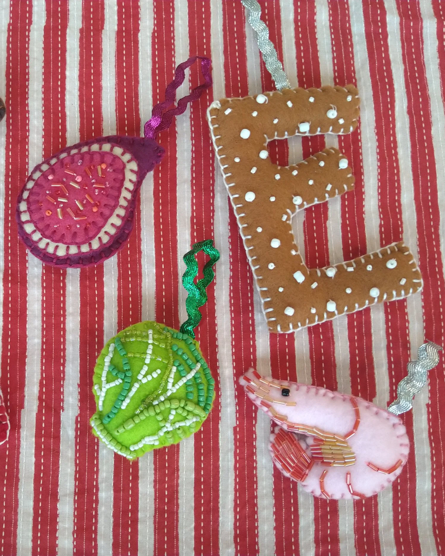 Colorful, decorated handmade felt and fabric Christmas ornaments in the shape of a pink fig, a prawn, and a gingerbread letter, and a brussel sprout with colorful beads and trim, on a red and cream striped fabric background.