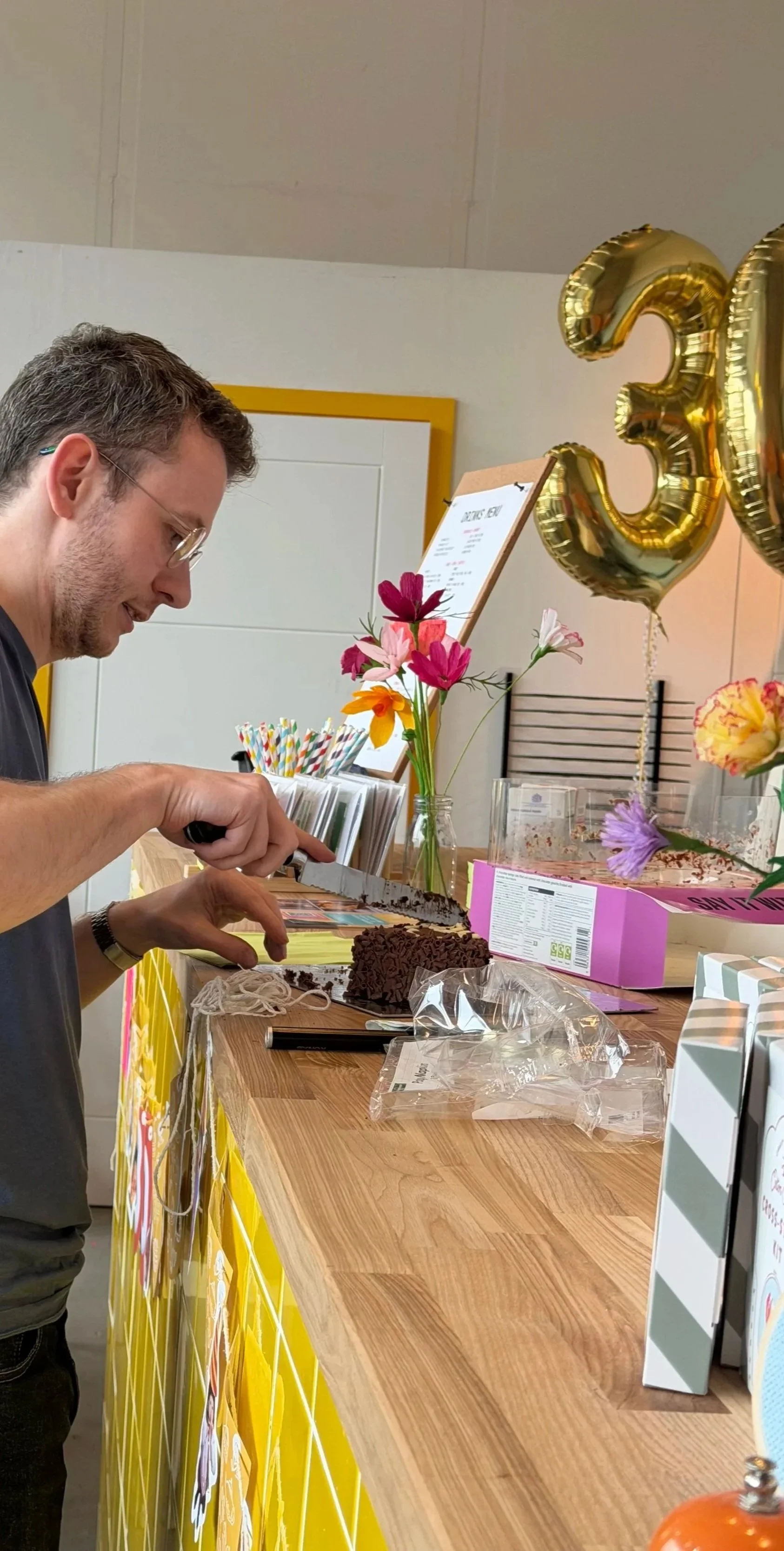 Man slicing a cake at a birthday celebration at Something Good Workshops with gold balloons displaying the number 30, colorful flowers, and party decor.