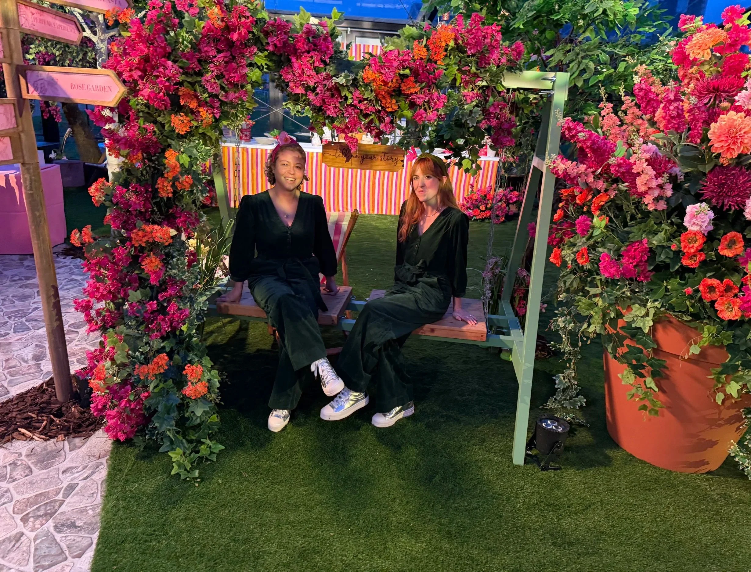 Two women sitting on a swing surrounded by pink, red, and orange flowers and green foliage at a garden or outdoor event during evening or night.