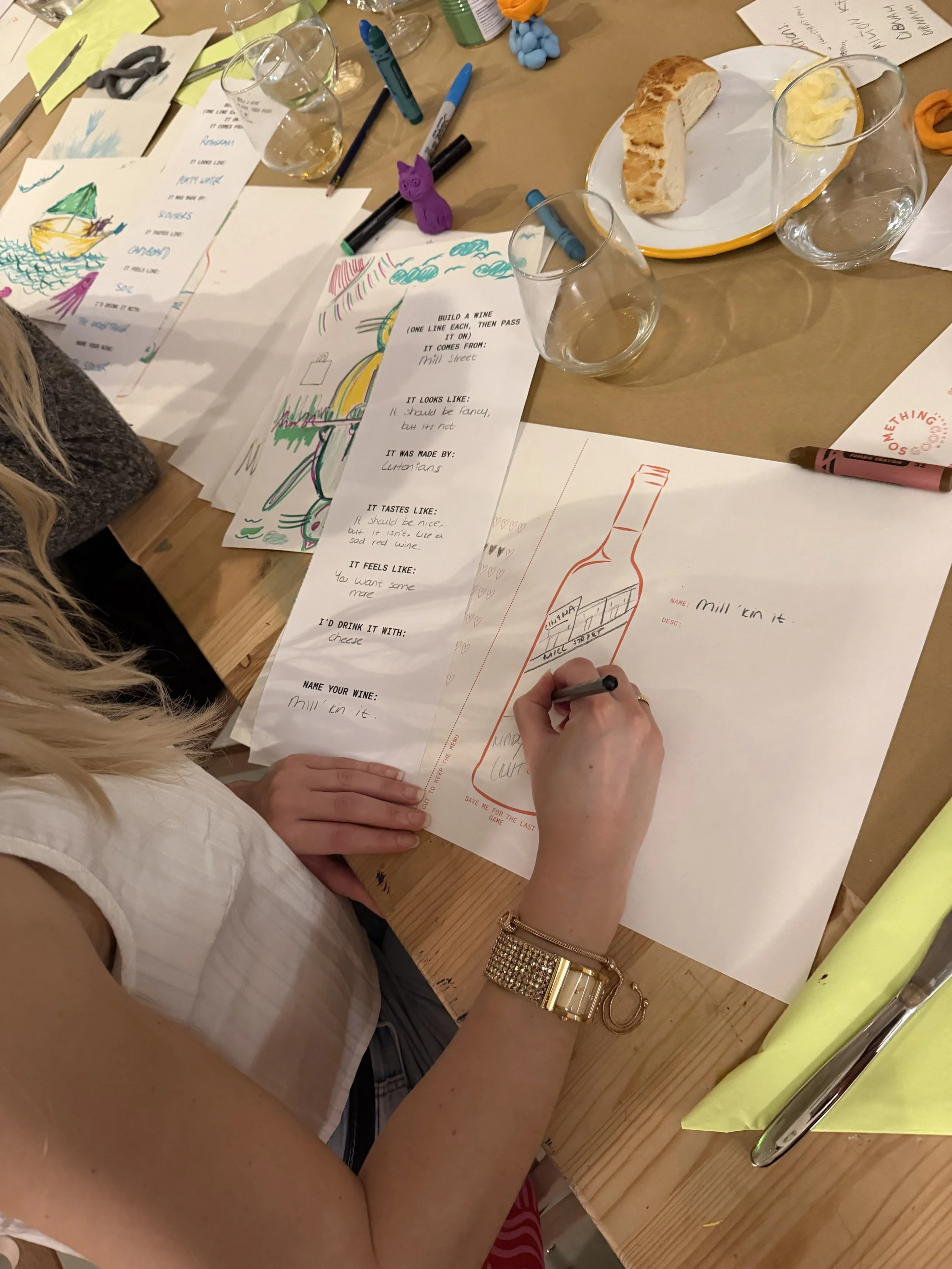 A person with long blonde hair is sitting at a wooden table, drawing and coloring on printed sheets of paper with sketches and instructions about wine. The table has various art supplies, including markers and pens, and some snacks like bread. There are empty wine glasses on the table, and a gold wristwatch is visible on the person's wrist.