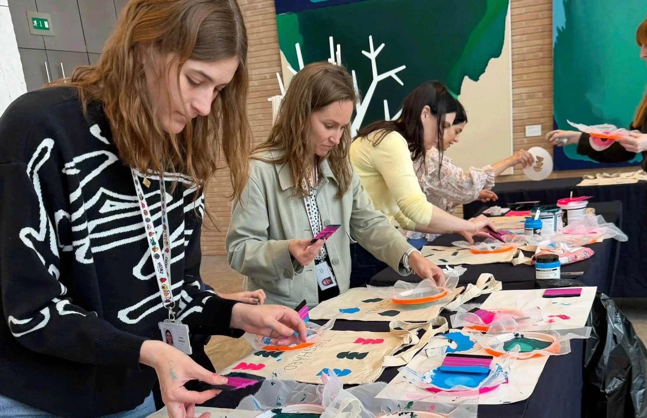 Women engaged in arts and crafts activity at a table, with various supplies, in an indoor office setting with abstract green and yellow art on the walls.