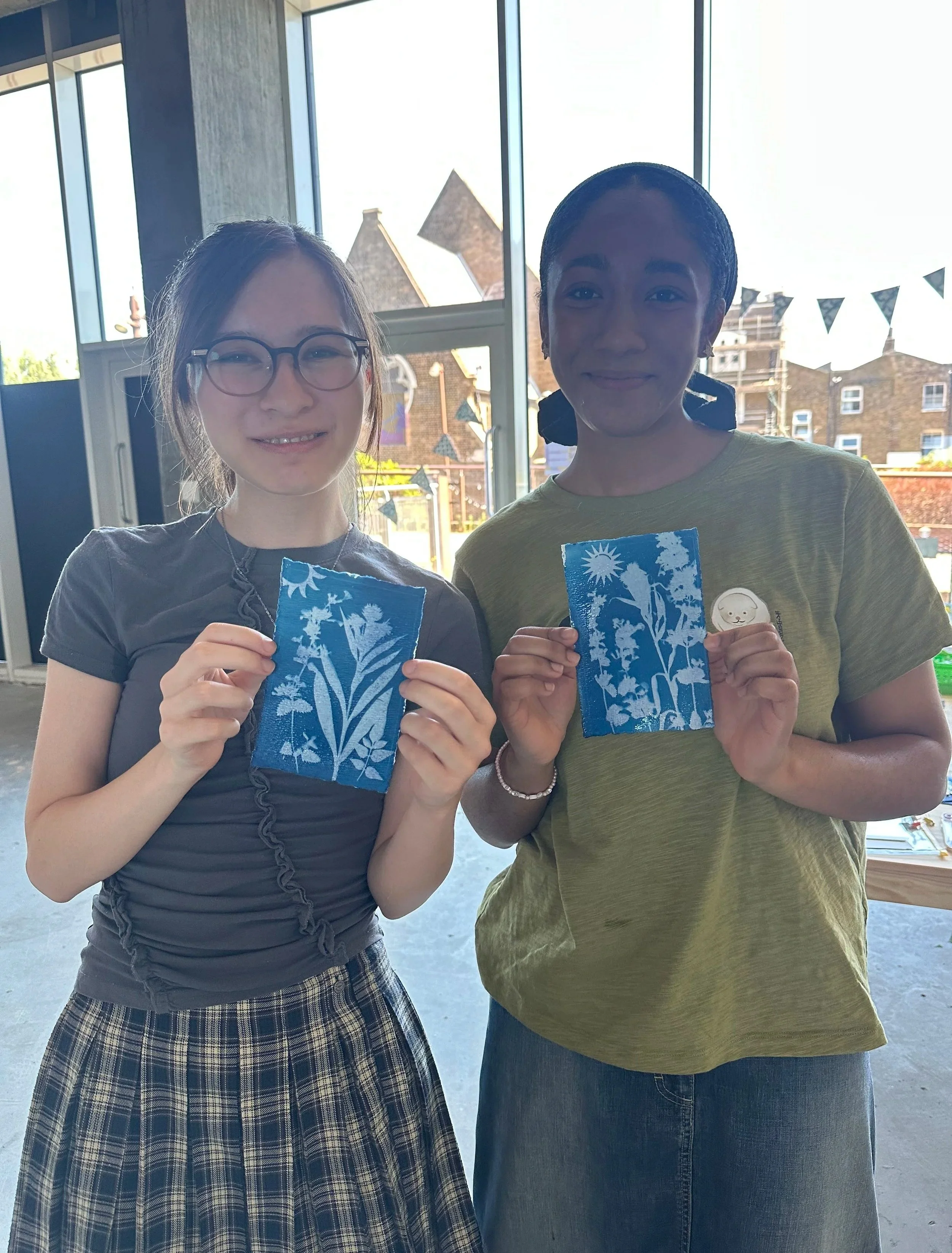 Two women standing indoors near large windows, holding blue pieces of paper with white floral and sun patterns, smiling at the camera, with outdoor buildings visible through the windows at a cyanotype workshop in Deptford, London.