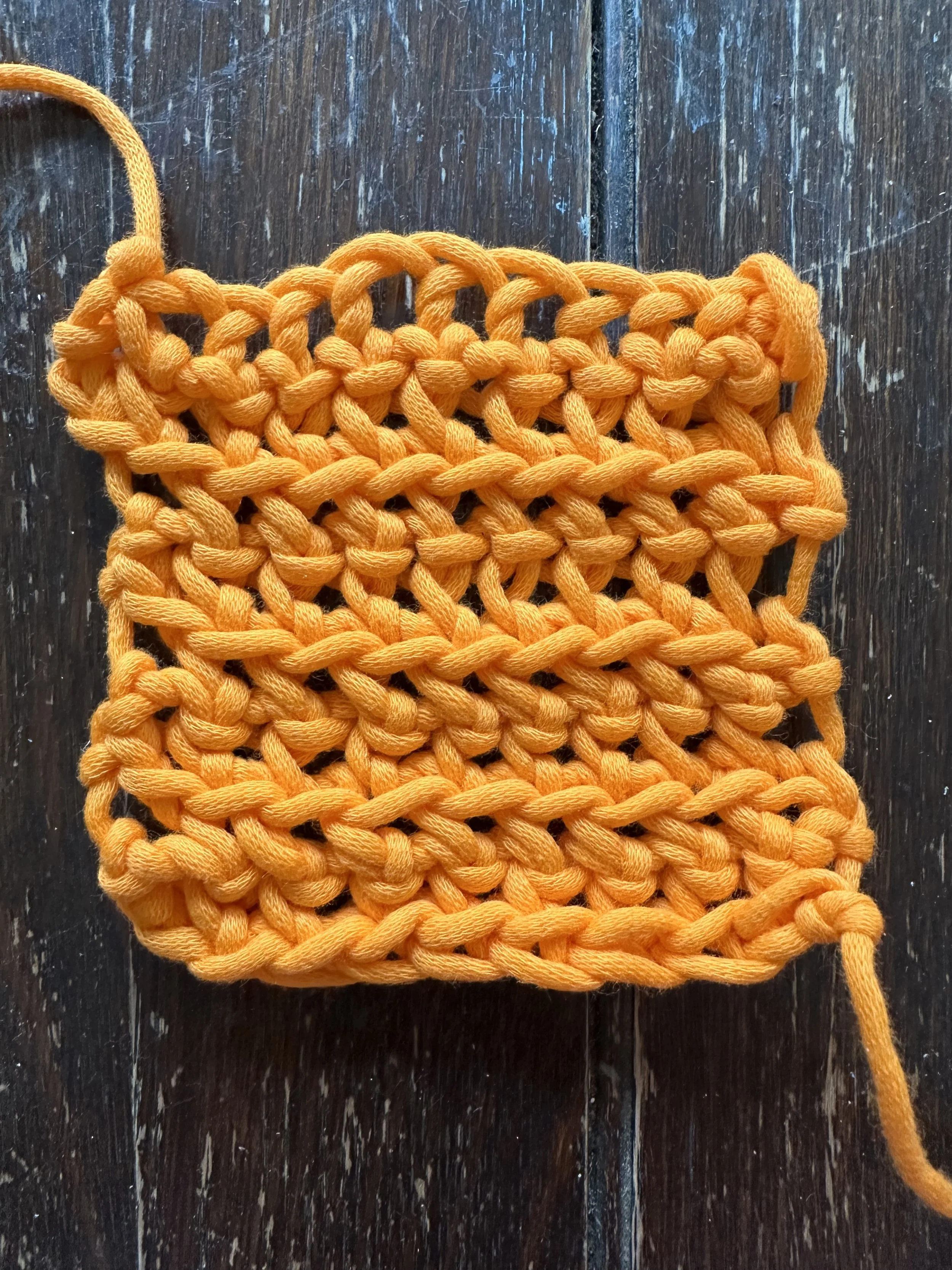 A close-up of an orange crochet square with chunky stitches on a dark wooden surface.
