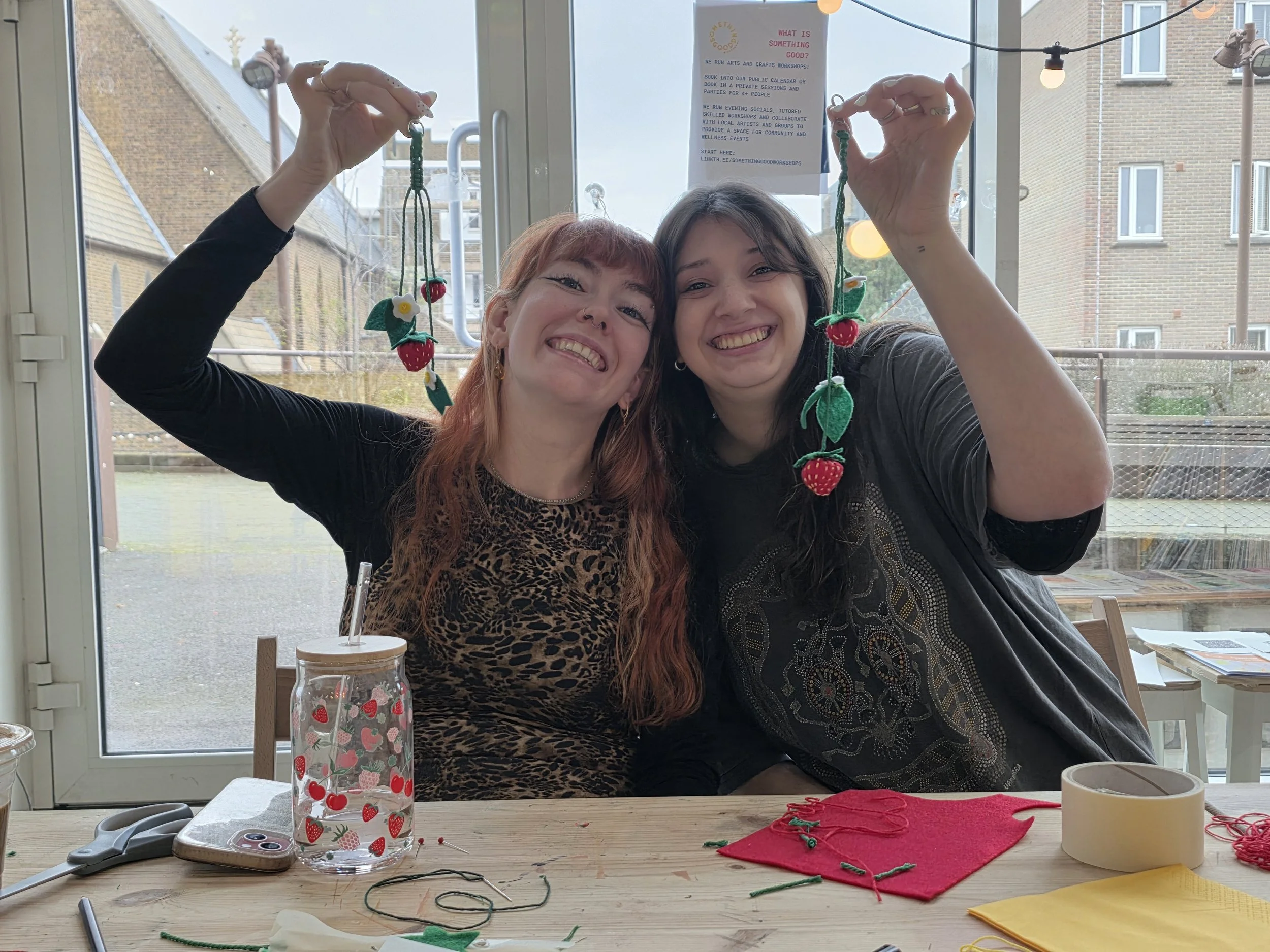 Two women smiling and holding up sewing projects with strawberries and leaves, sitting at a table with crafting supplies, in a bright room with large windows.