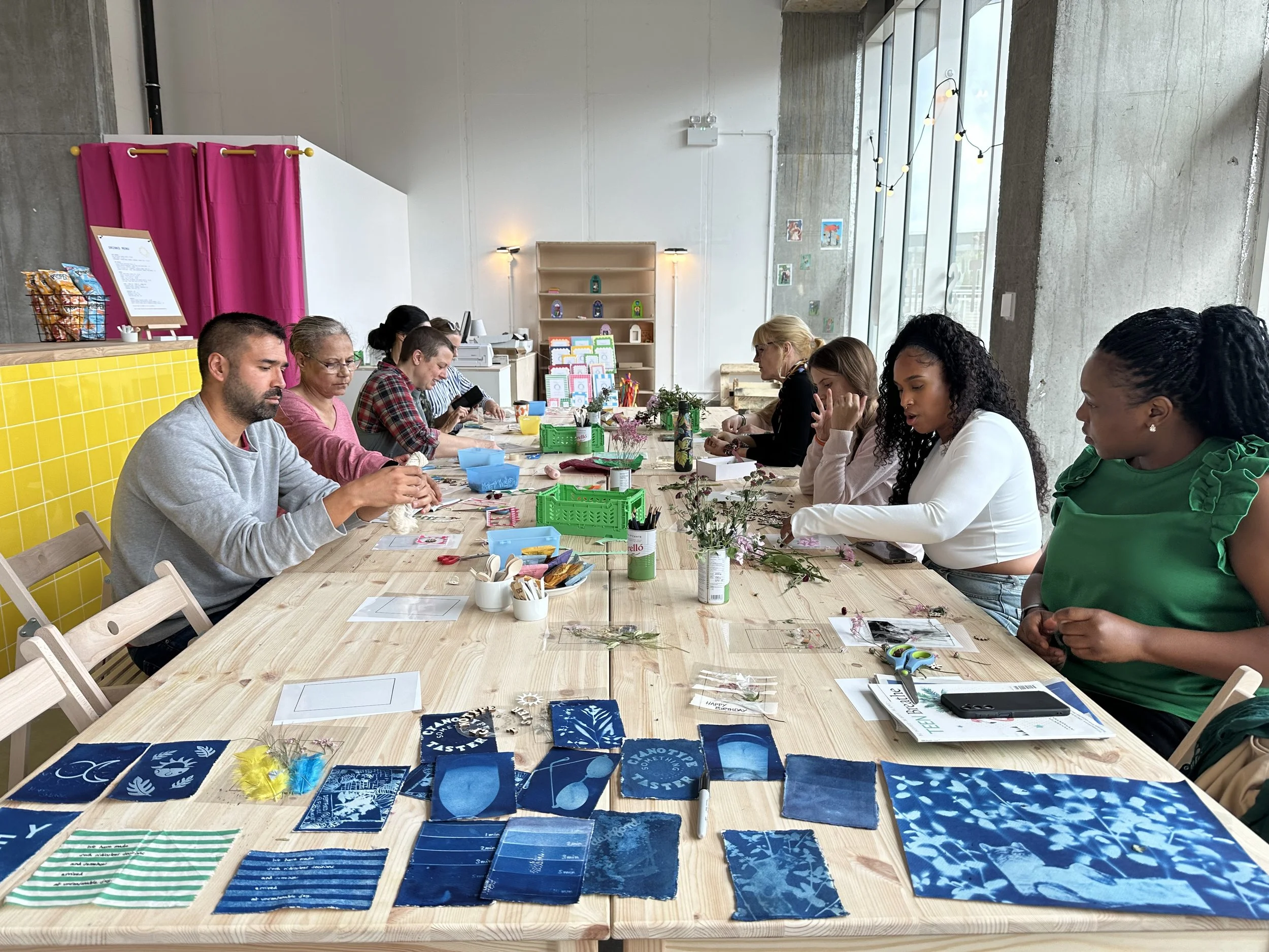 A group of people sitting at a long wooden table engaged in a craft activity, with craft supplies and fabric swatches displayed on the table, in a bright room with large windows and string lights.