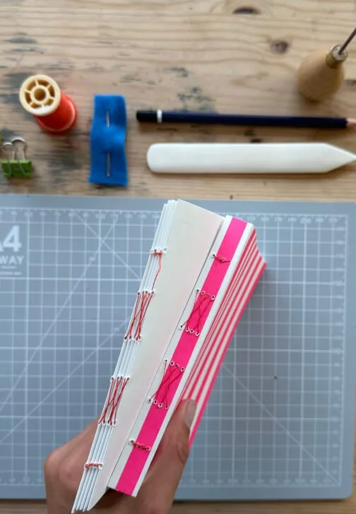 A person holding a small french link stitch book with pink and white striped pages, with chessboard patterns on the cover, surrounded by sewing and craft tools on a work surface at a bookbinding workshop in London.