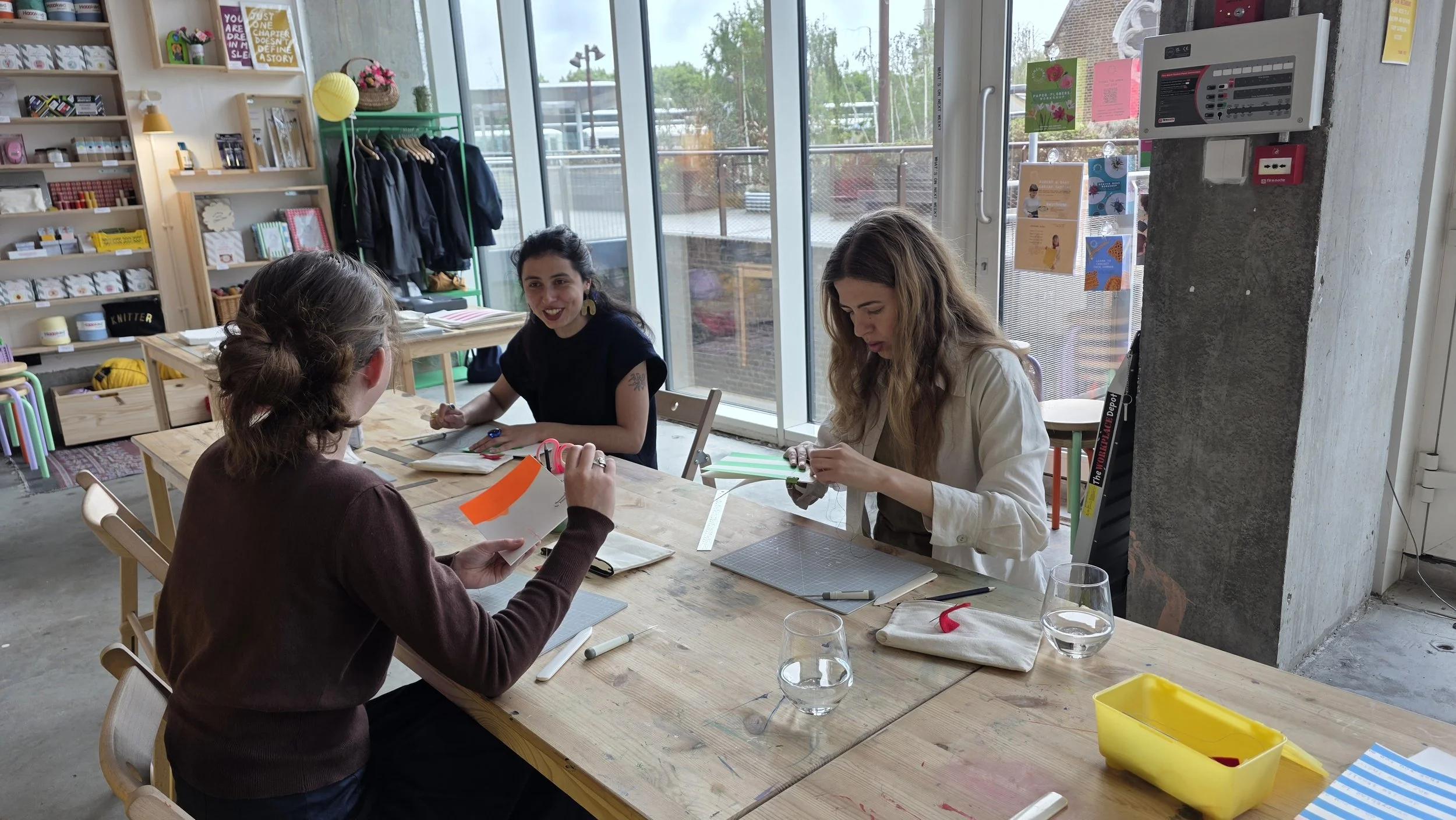 Three women sitting around a wooden table engaging in a creative activity in a bright, modern craft or coworking space, with shelves of supplies and large glass windows in the background.