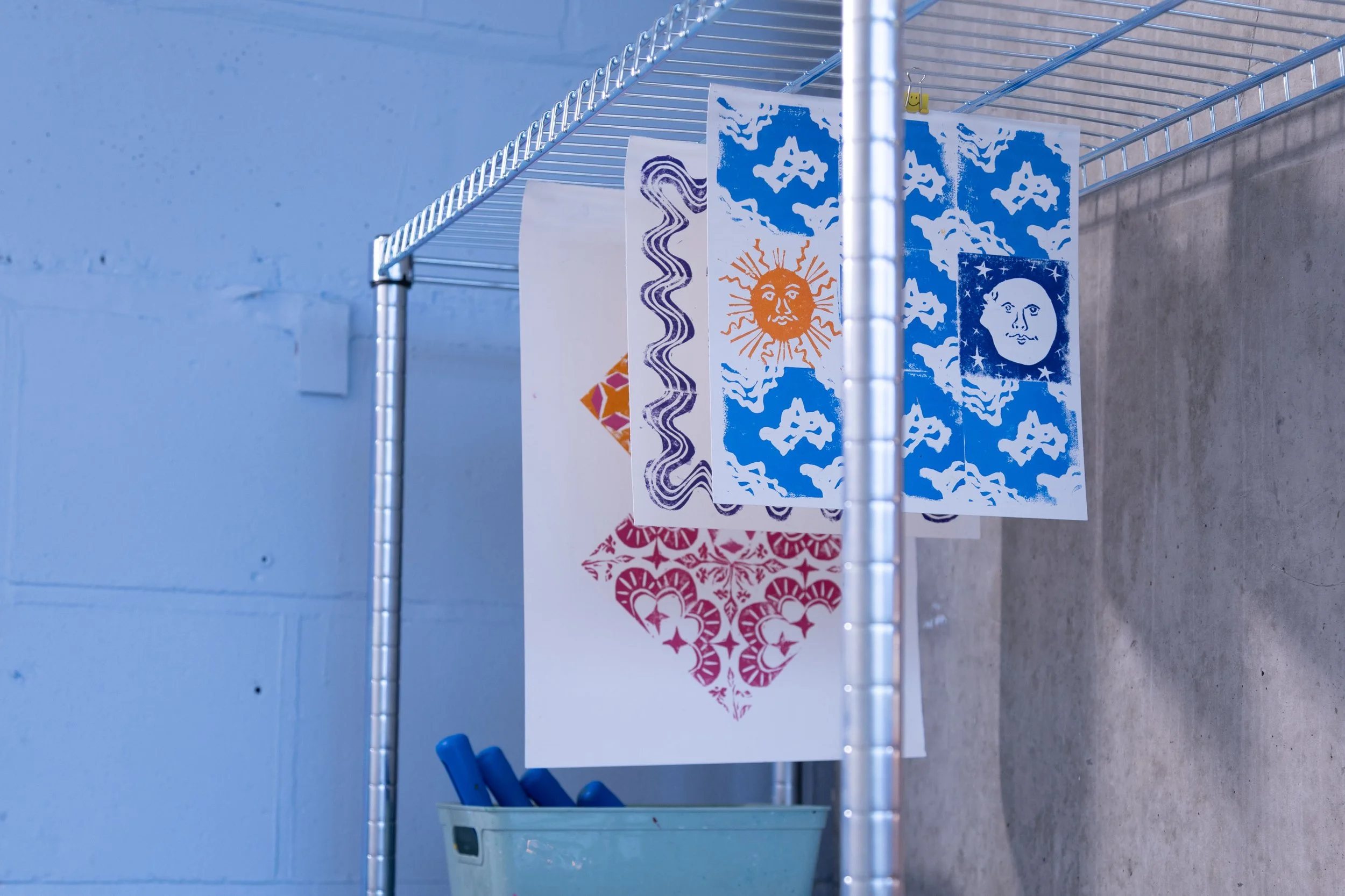 Colorful decorative paper or artwork hanging on a metal shelf against a blue wall, with a plastic bin containing blue markers or pens below.