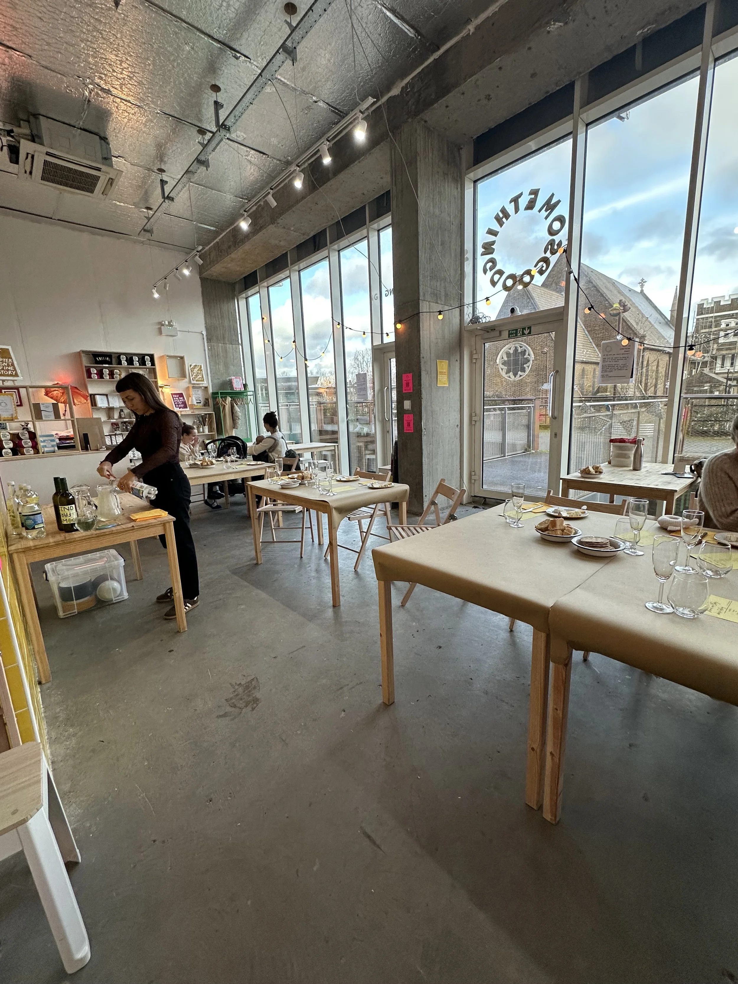 Interior of a modern restaurant with large windows, decorated with string lights, set with tables, plates, and glasses, and a woman preparing food.