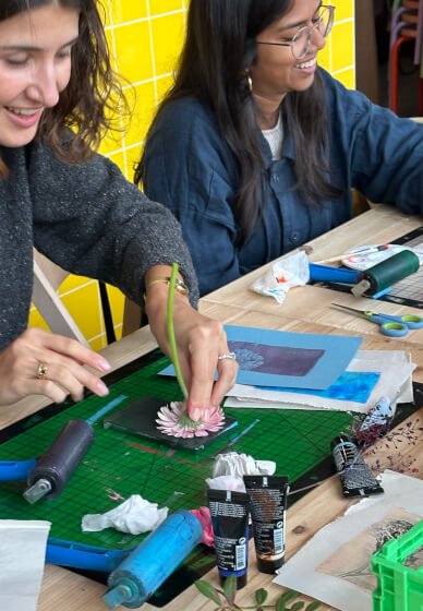 Two women working on a gelli plate printing craft project at a table with art supplies, including glue, scissors, and paper.