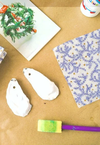 Decorative holiday items and craft supplies on a table, including a small decorated Christmas tree on a book, blue and white holiday paper, two oyster shells, a green and purple paintbrush, and a container of glue.