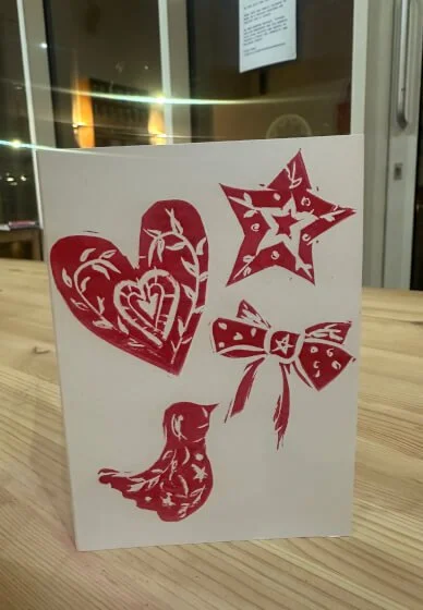 A white card featuring lino printed red decorative stamps of a heart, star, bird, and bow, placed on a wooden surface near a window.