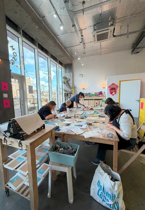 People engaging in crafting or artwork at a long table in a well-lit urban studio with large windows, bookshelves, and colorful decor.