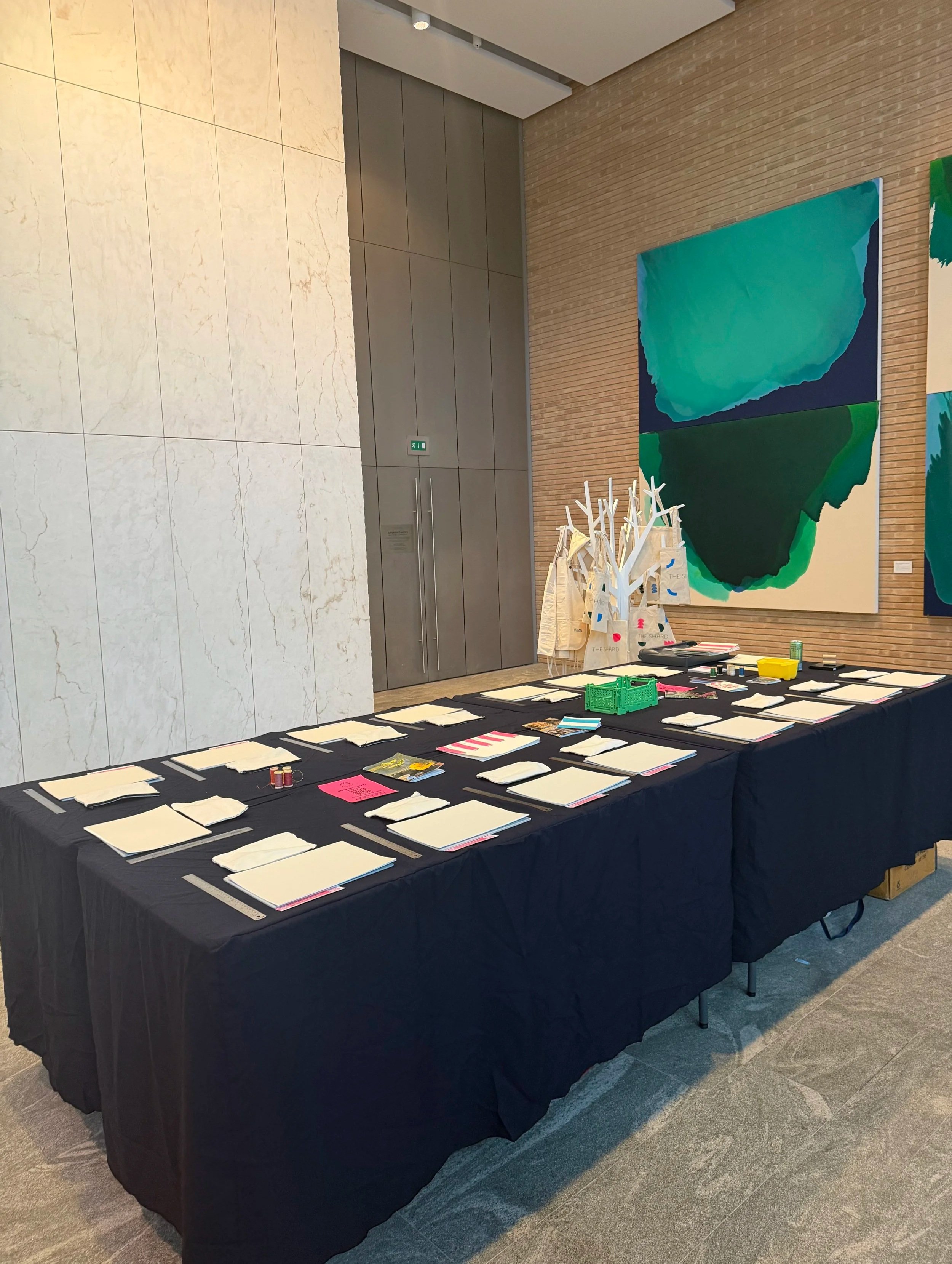 A large table covered with a black tablecloth displays multiple stacks of paper, writing utensils, and small containers in an indoor setting with art on the walls and an elevator in the background.