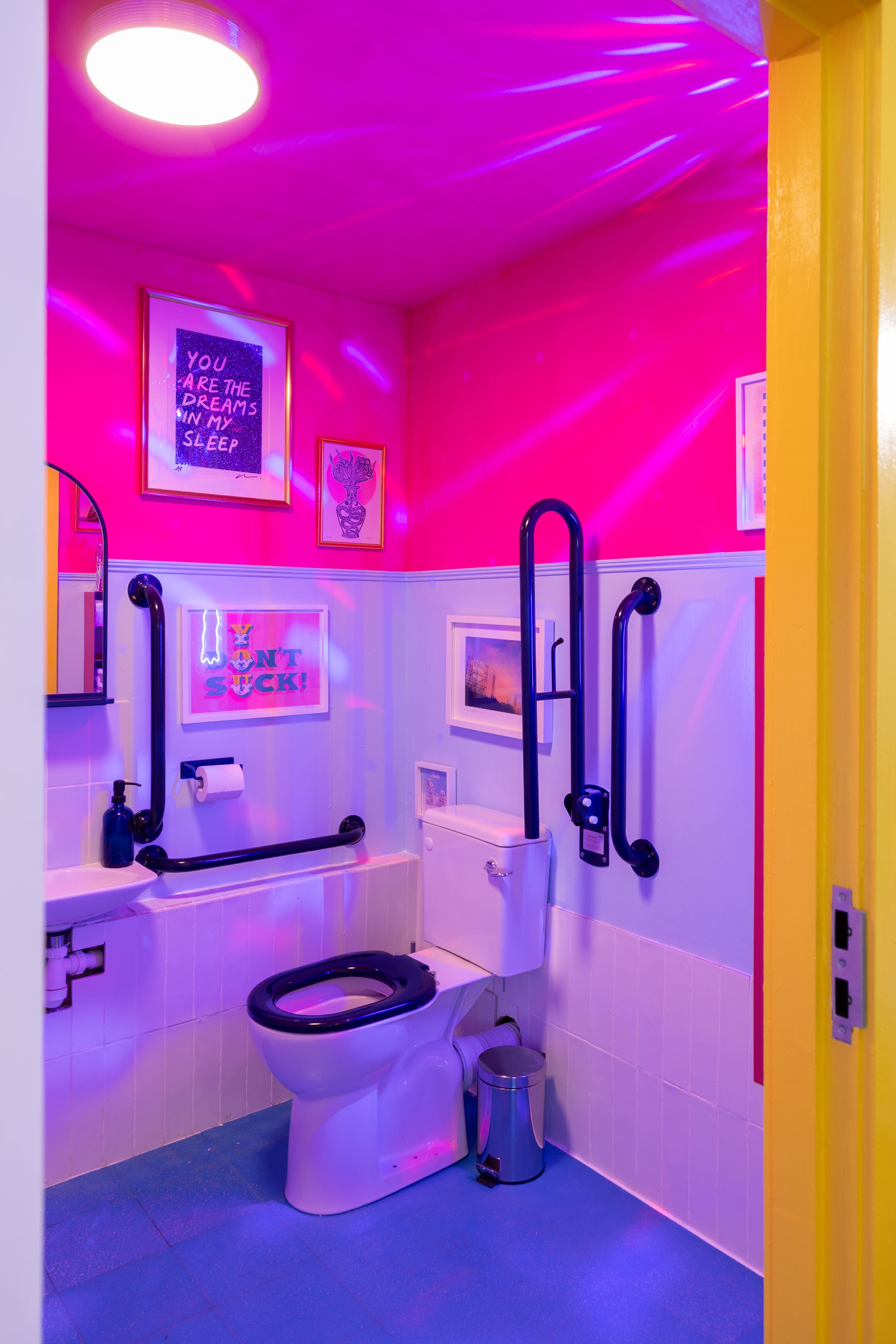 A bathroom with a pink wall, blue flooring, and various framed art. Purple and blue lights create a colorful atmosphere. The bathroom has a toilet with black and white seat, black handrails, a small trash can, and a toilet paper holder.