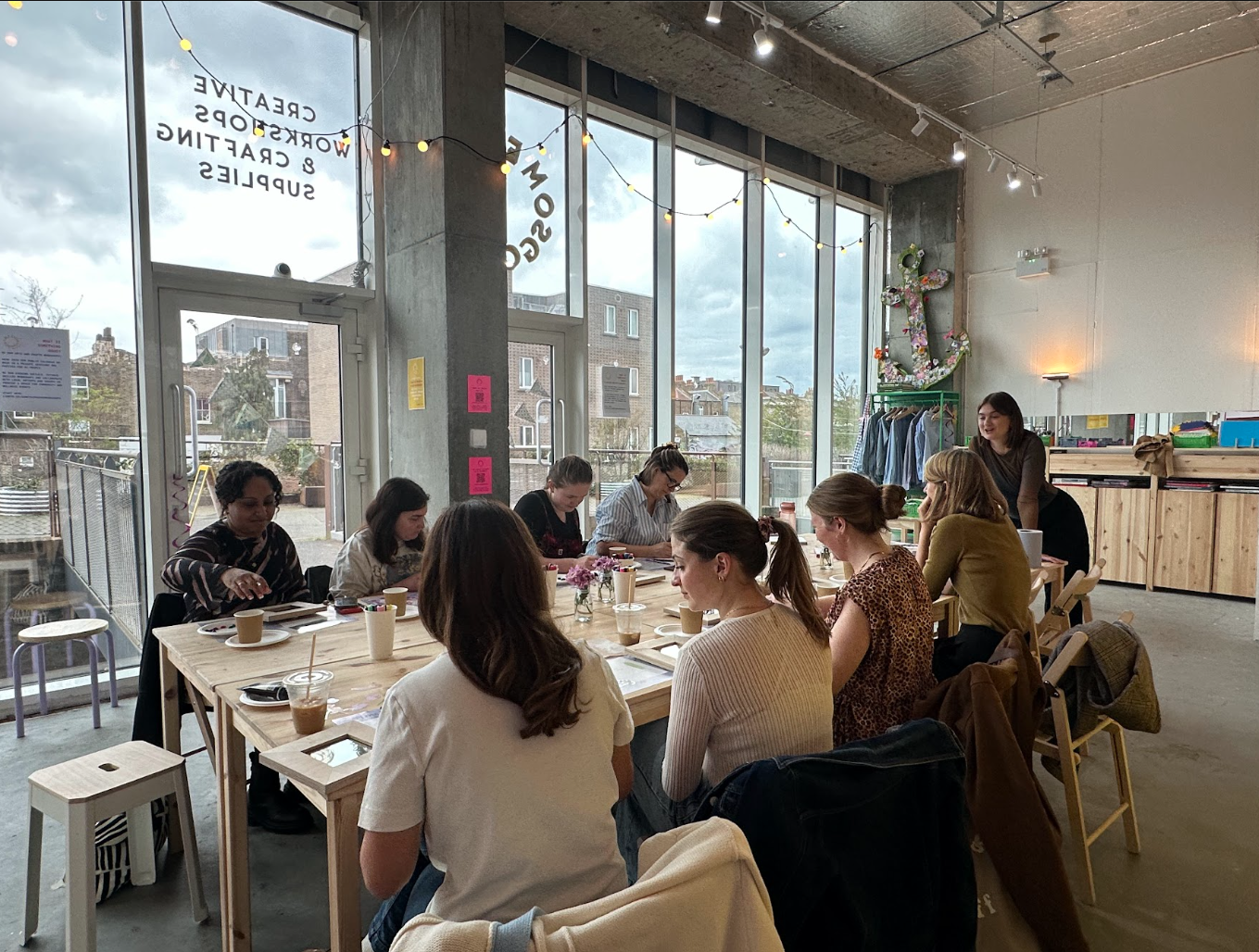 A group of women sitting around a long wooden table in a modern, well-lit with large windows, during daytime. There are drinks, flowers, and notebooks on the table. A woman is standing and smiling at the side. 