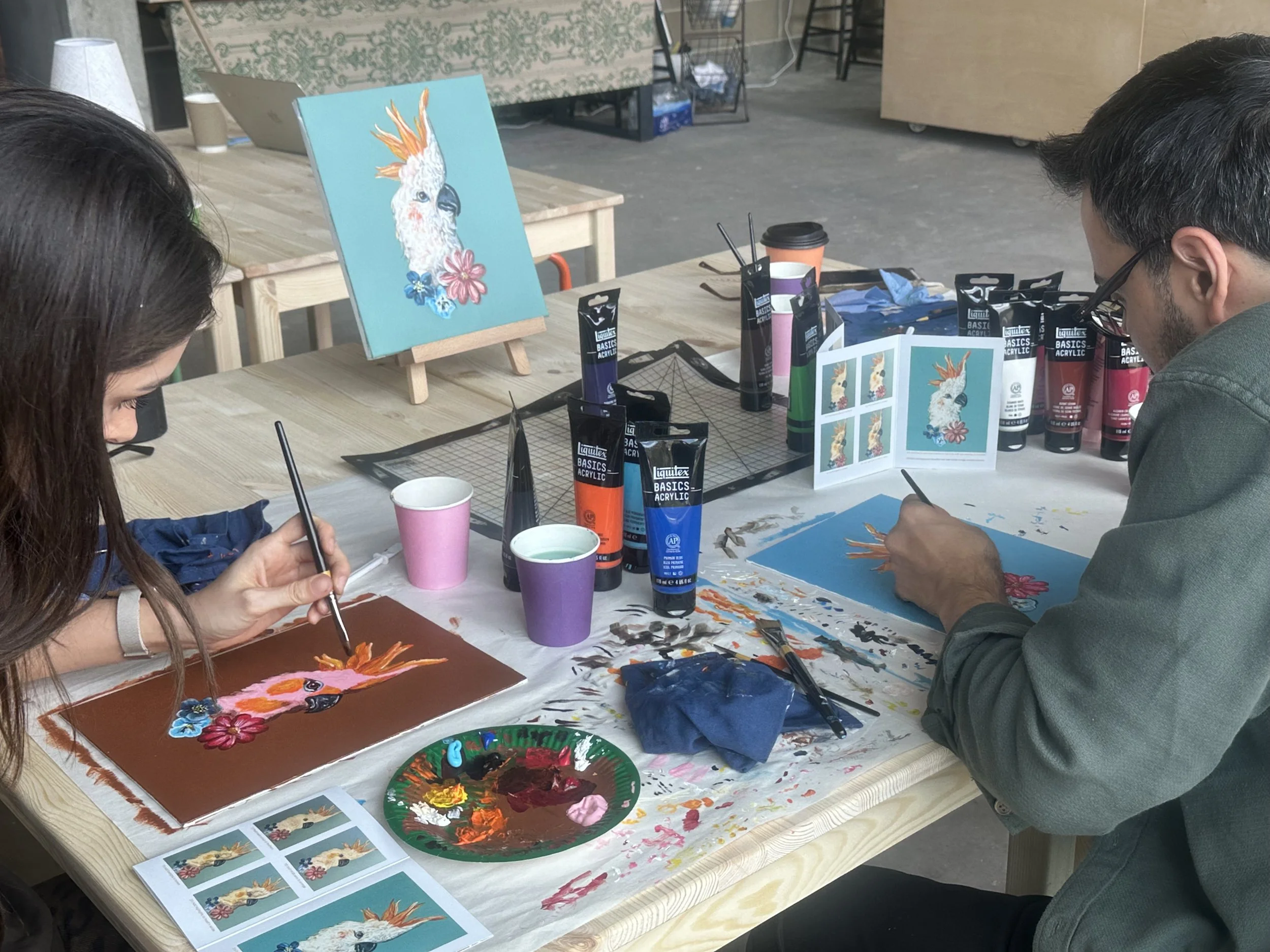 Two people painting a colorful parrot with flowers on small canvases at a wooden table, with paint bottles, brushes, and reference images on the table.