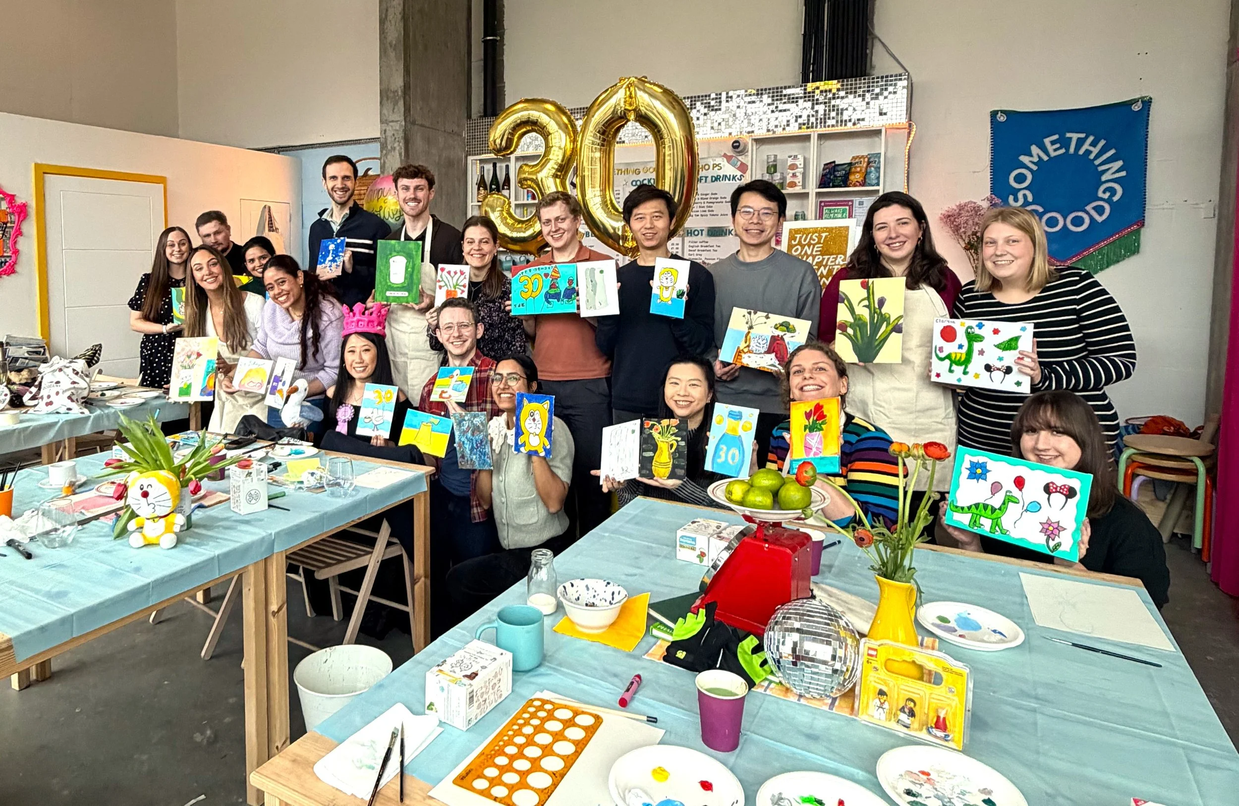 Group of people at a painting workshop in London holding up their completed artwork beneath large gold ‘30’ balloons.