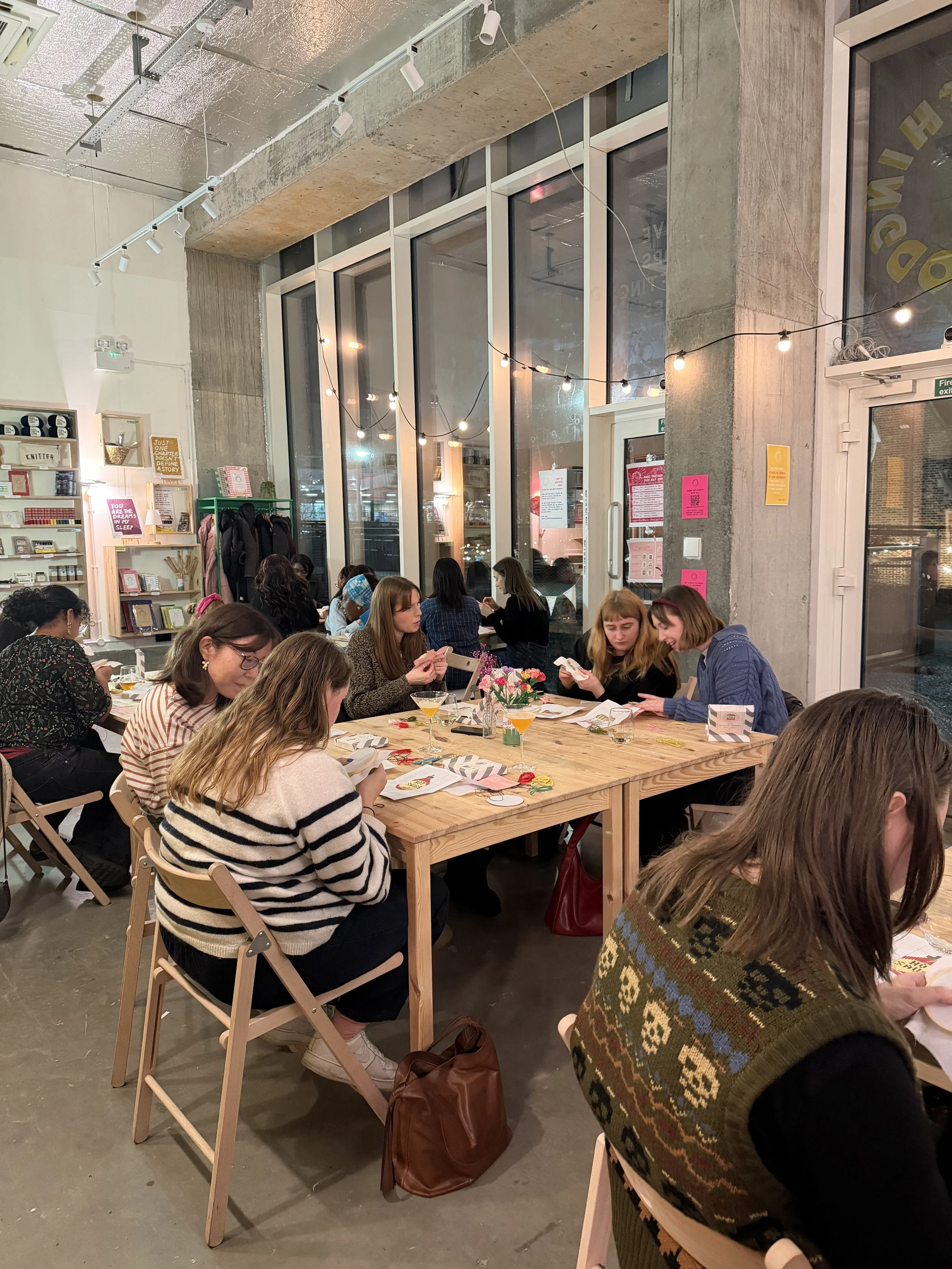 A group of women sitting around a large wooden table, engaged in a cross stitch workshop, inside a modern, well-lit craft workshop with large windows, string lights, and bookshelves.