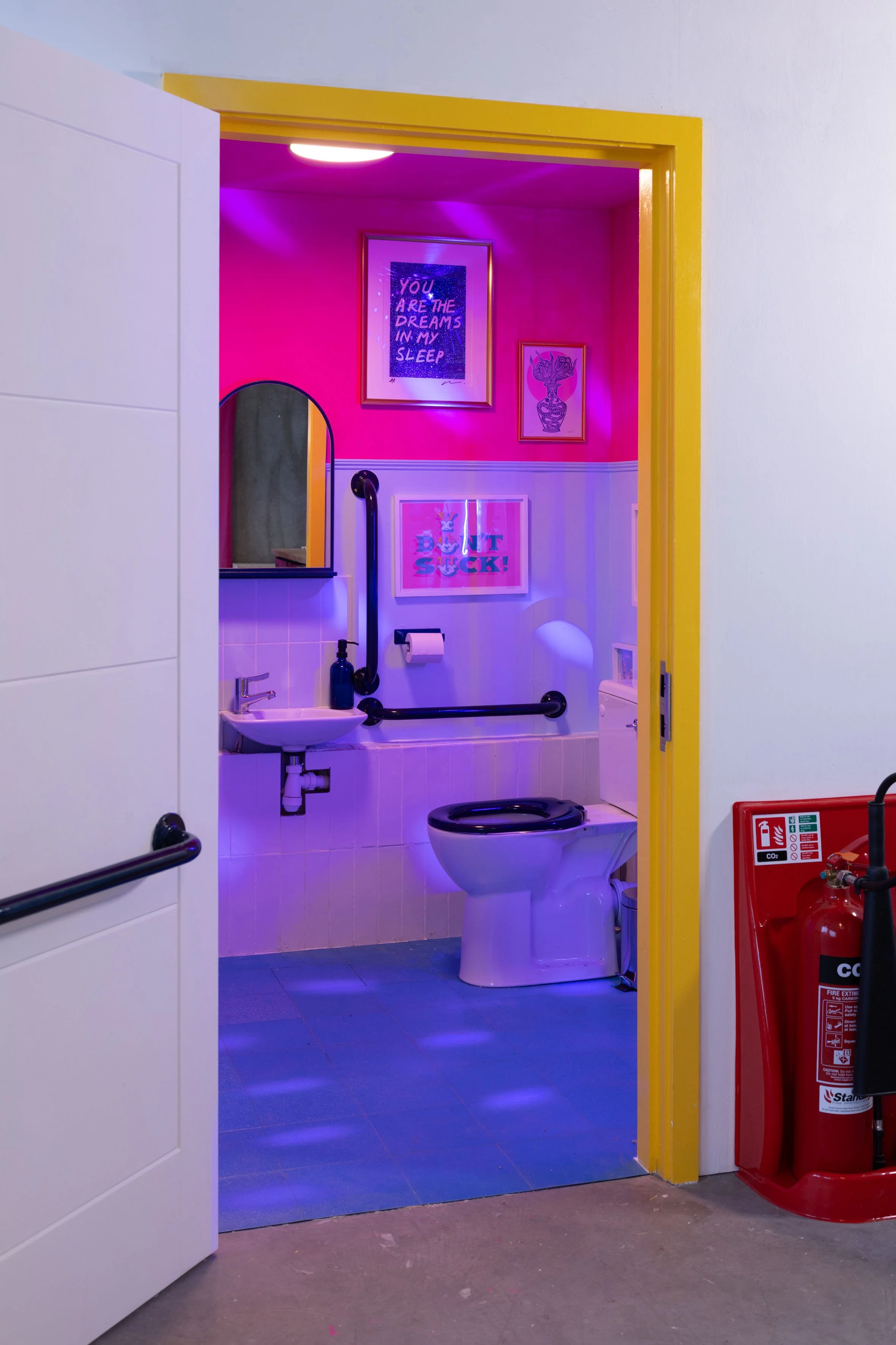 Colorfully decorated bathroom with pink and purple lighting, framed wall art, and a yellow door frame, featuring a toilet, a small sink, mirrored cabinet, and handrails.