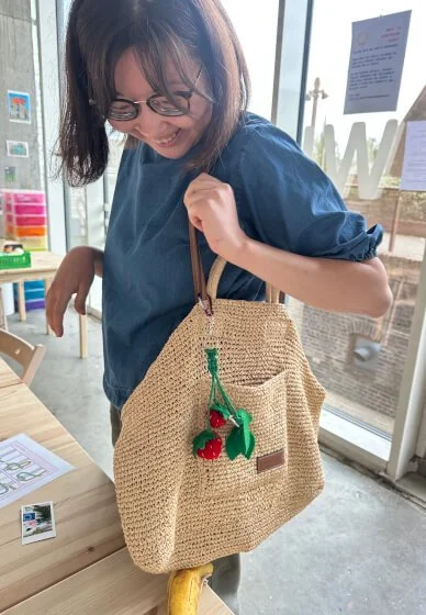 A woman with glasses wearing a blue shirt and holding a woven beige handbag decorated with a handmade strawberry bag charm, standing inside a store or cafe near large windows.