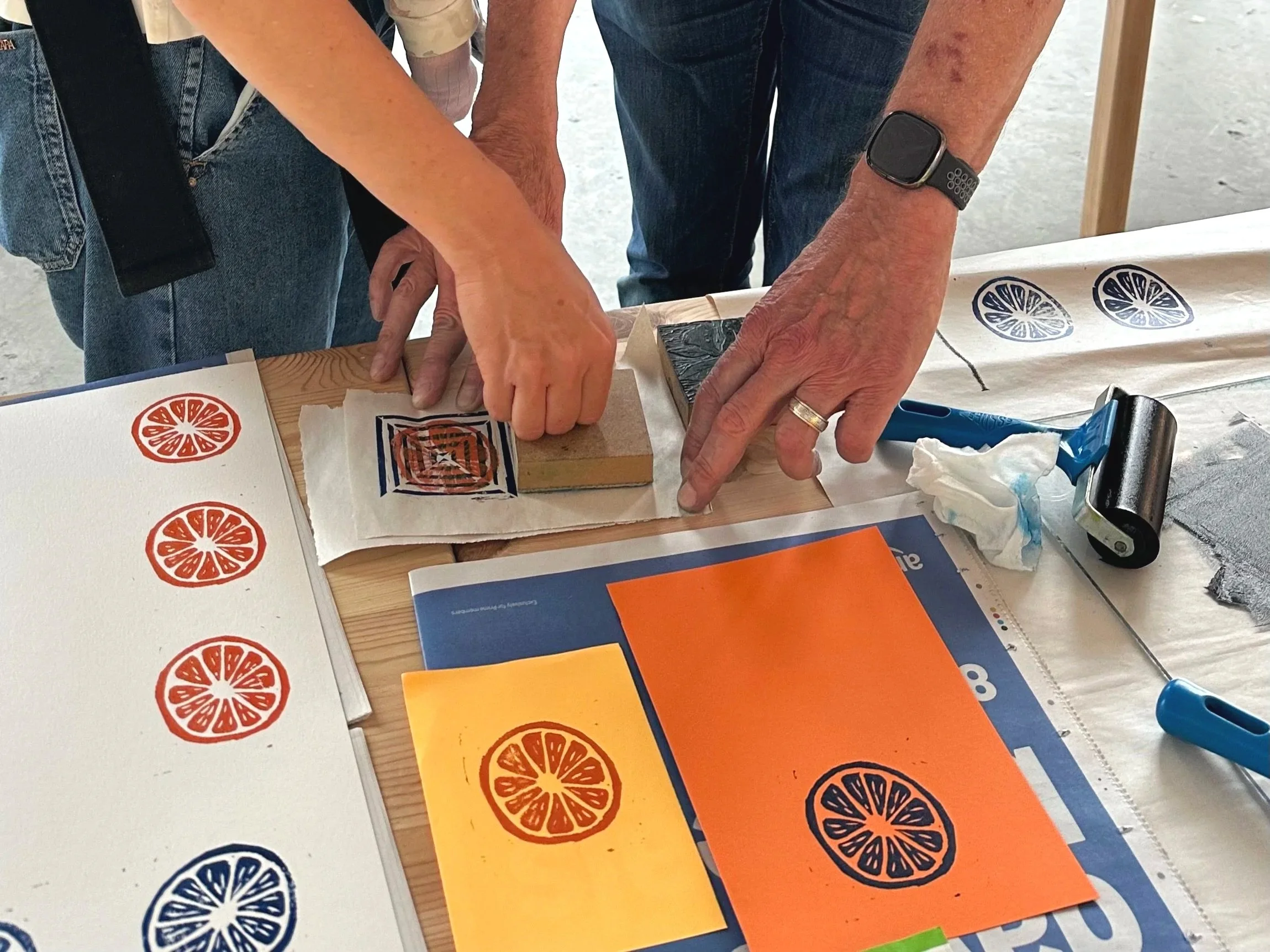 Close-up of two people lino printing orange and black citrus fruit designs onto paper at a table, surrounded by study materials and art supplies.