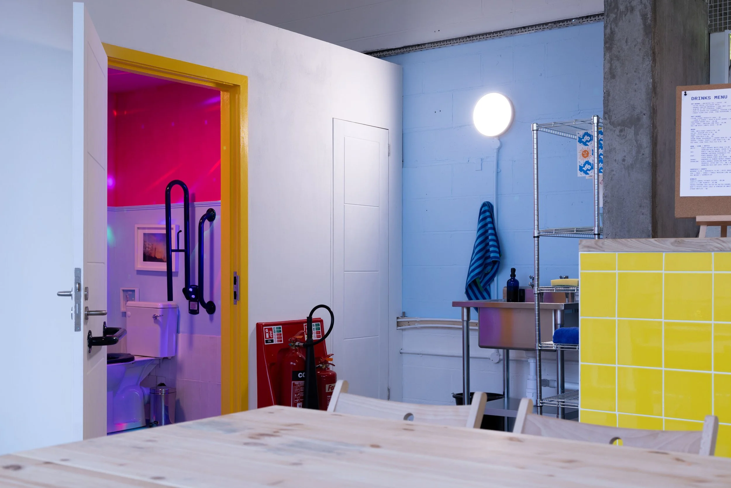Interior of a room with a yellow-tiled counter, a sink, and shelving on a blue wall, a towel, a round light, a fire extinguisher, and a doorway with pink and purple lighting leading to a bathroom with a toilet and black wall-mounted grab bars.